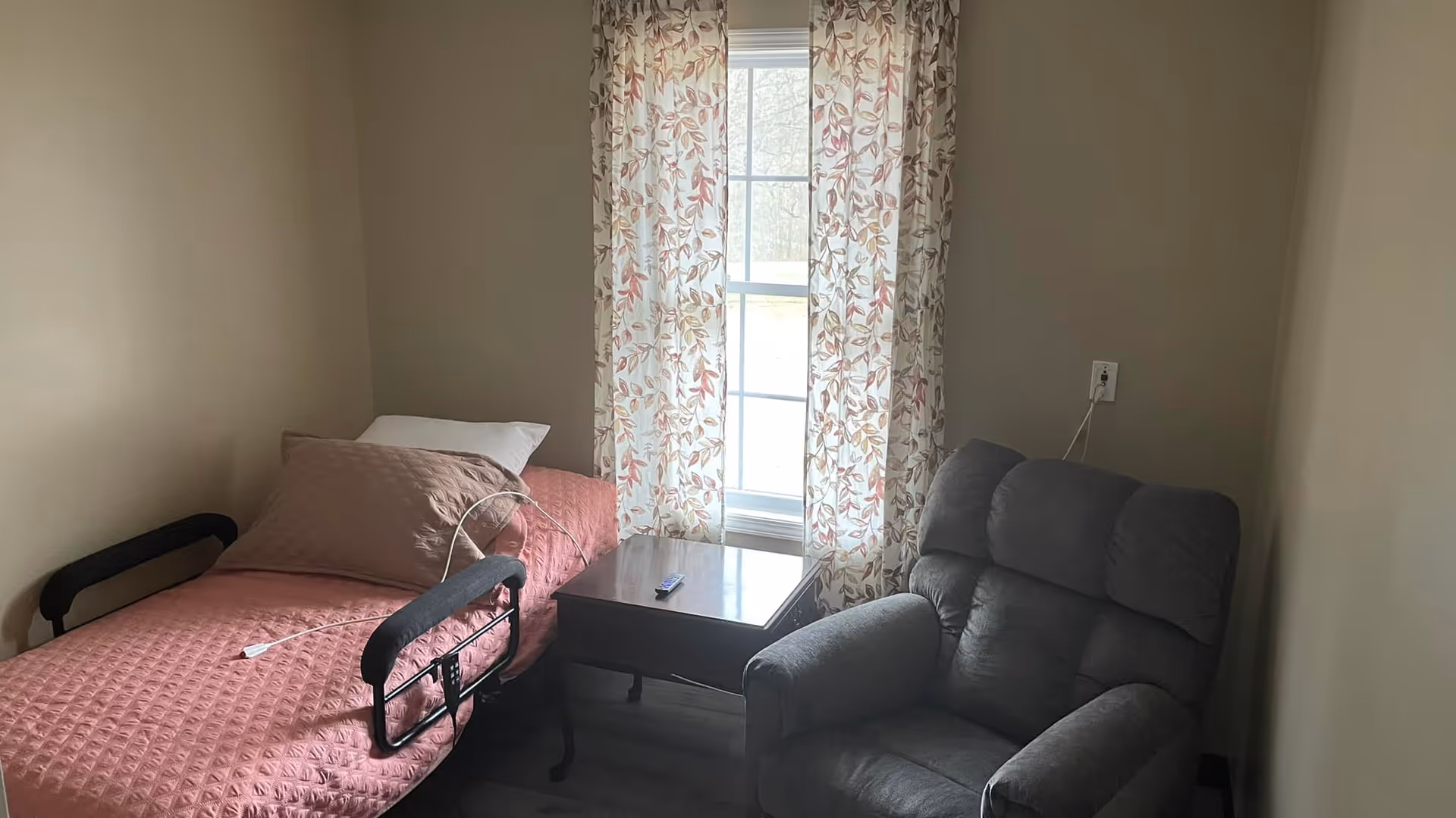Cozy bedroom with pink quilt bed, floral curtains, window, and gray recliner.