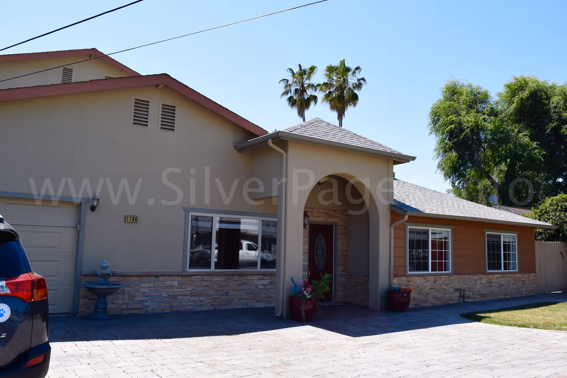 Single-story senior residence with arched entrance, beige walls, large windows, palm trees, and driveway.