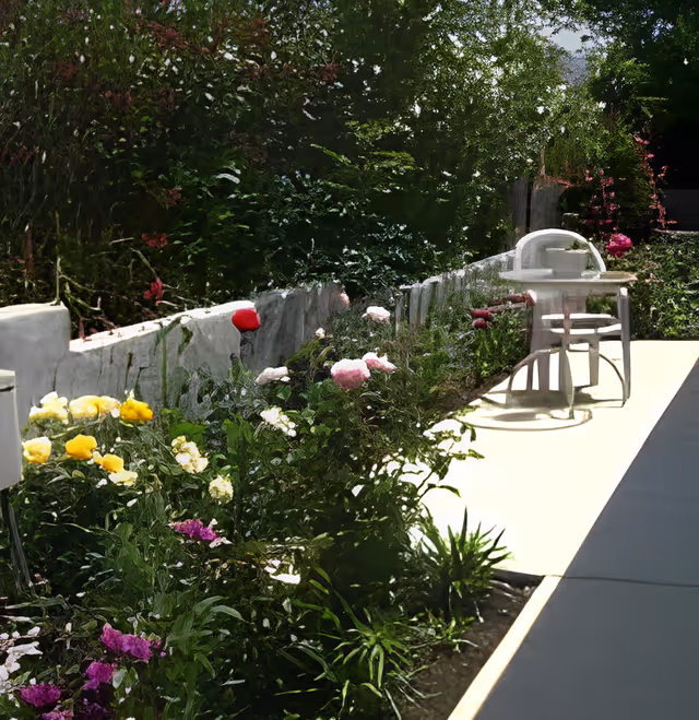 Sunlit outdoor patio with a narrow walkway, flowerbeds along a low stone wall, and a small table and chairs.
