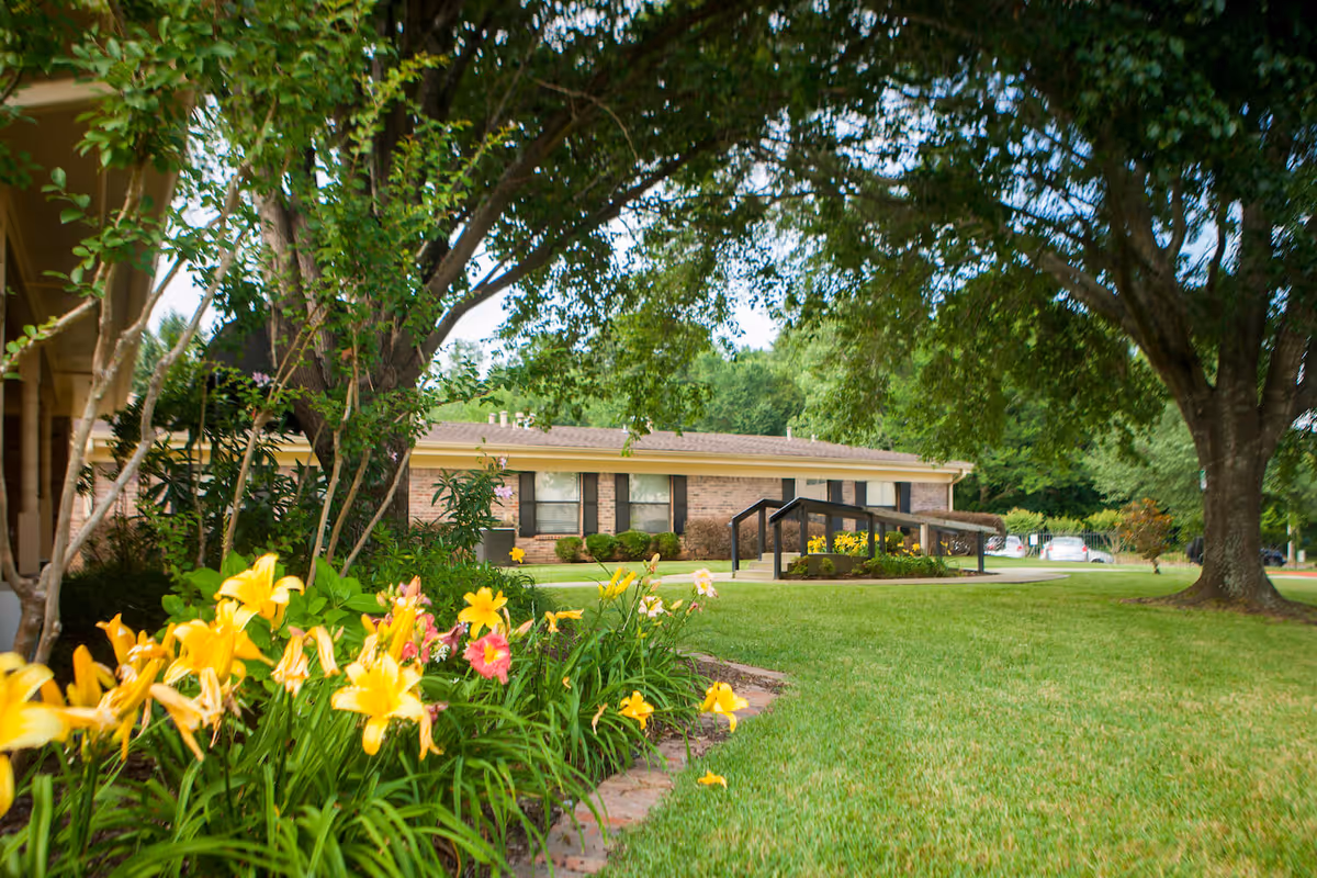 A well-maintained outdoor garden area at Briarcliff Health Center featuring a lush green lawn, colorful yellow and pink flowers, large trees providing shade, and a brick building with windows and a ramp in the background.
