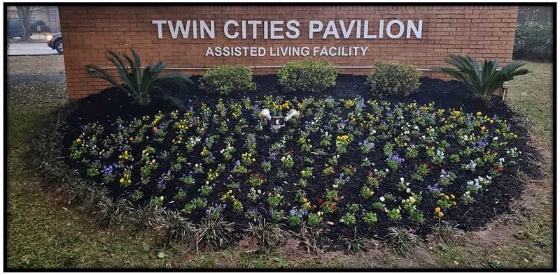 Brick entrance sign reading "Twin Cities Pavilion Assisted Living Facility" above a landscaped flower bed with rows of small flowering plants.