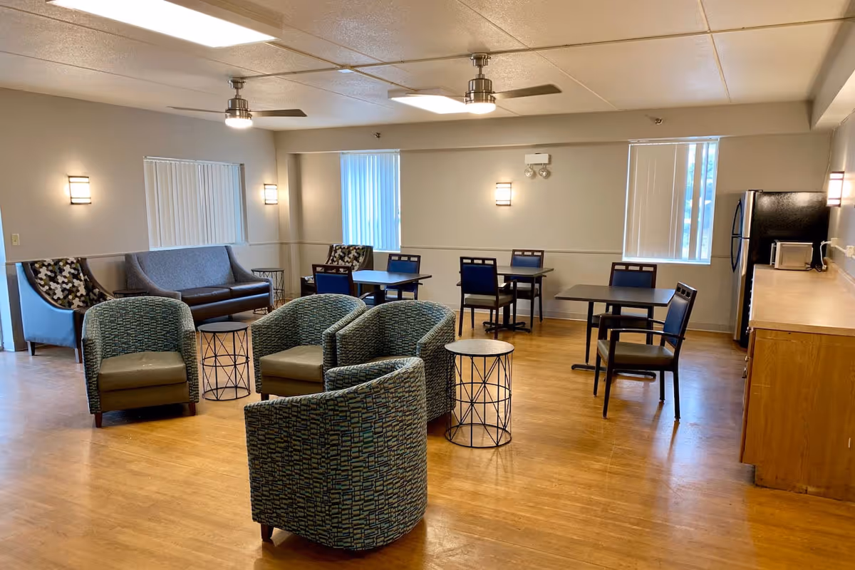 Open communal lounge with grouped armchairs, sofas, dining tables and a kitchenette in a senior living facility.