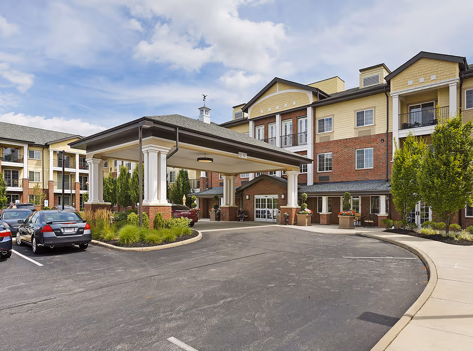 Front entrance of a multi-story senior living facility with a covered porte-cochere, parked cars, and landscaped grounds.