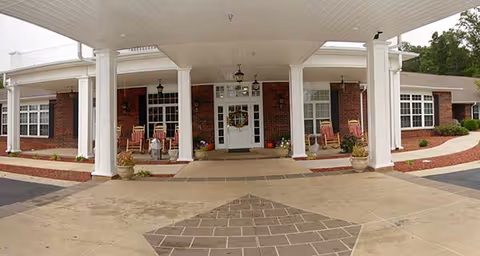 Covered entrance of a brick senior living facility with white columns, rocking chairs, potted plants, and double doors.