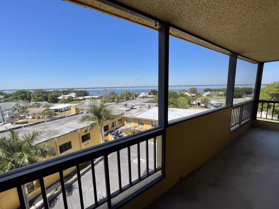 Balcony view over rooftops to a distant body of water beyond palm trees.