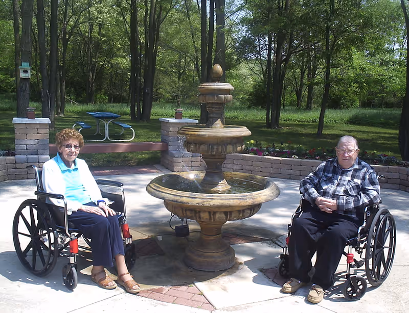An elderly woman and an elderly man sitting in wheelchairs on either side of a stone water fountain in an outdoor garden area with trees and a picnic table in the background.