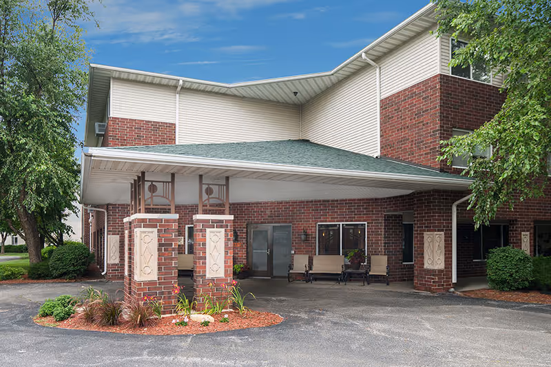 Exterior view of a two-story brick and siding building with a covered entrance supported by brick pillars. There are benches near the entrance and landscaping with bushes and trees around the building under a blue sky with some clouds.