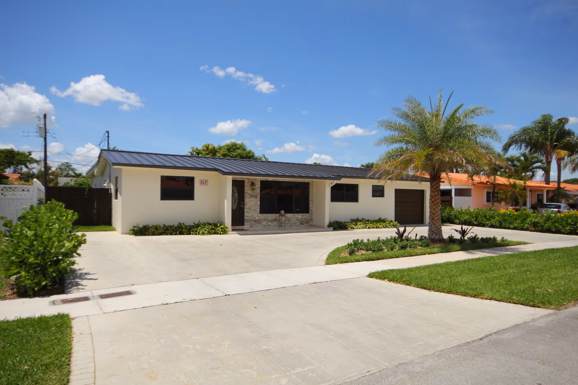 Single-story building with a modern design, white exterior walls, a dark metal roof, and a stone accent around the entrance door. There is a driveway and a small landscaped area with a palm tree and other plants in front of the building under a clear blue sky.
