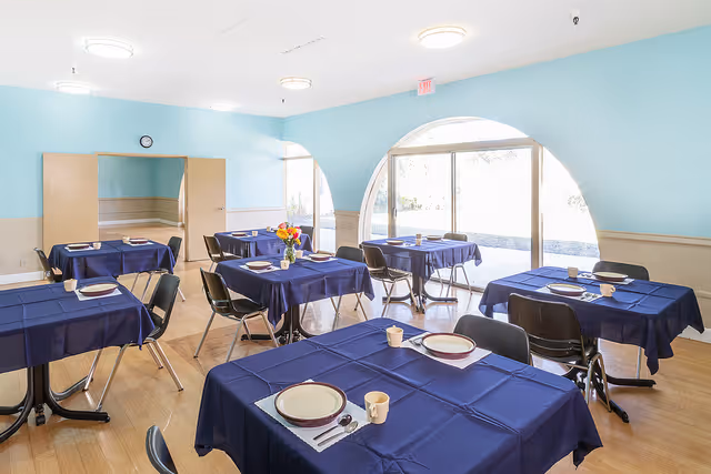 A bright dining room with multiple tables covered in dark blue tablecloths, each set with plates, cups, and utensils. The room has light blue walls, large arched windows letting in natural light, and wooden flooring. Several black chairs are arranged around the tables.