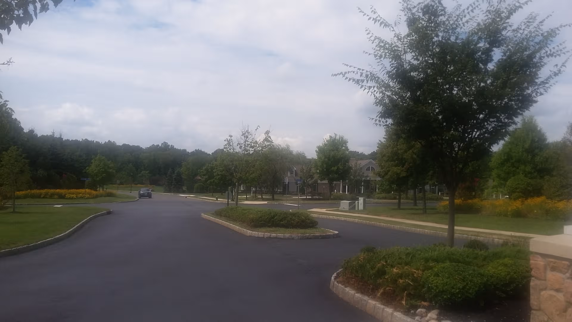 Wide roadway through a landscaped senior living campus with trees and a distant building visible.