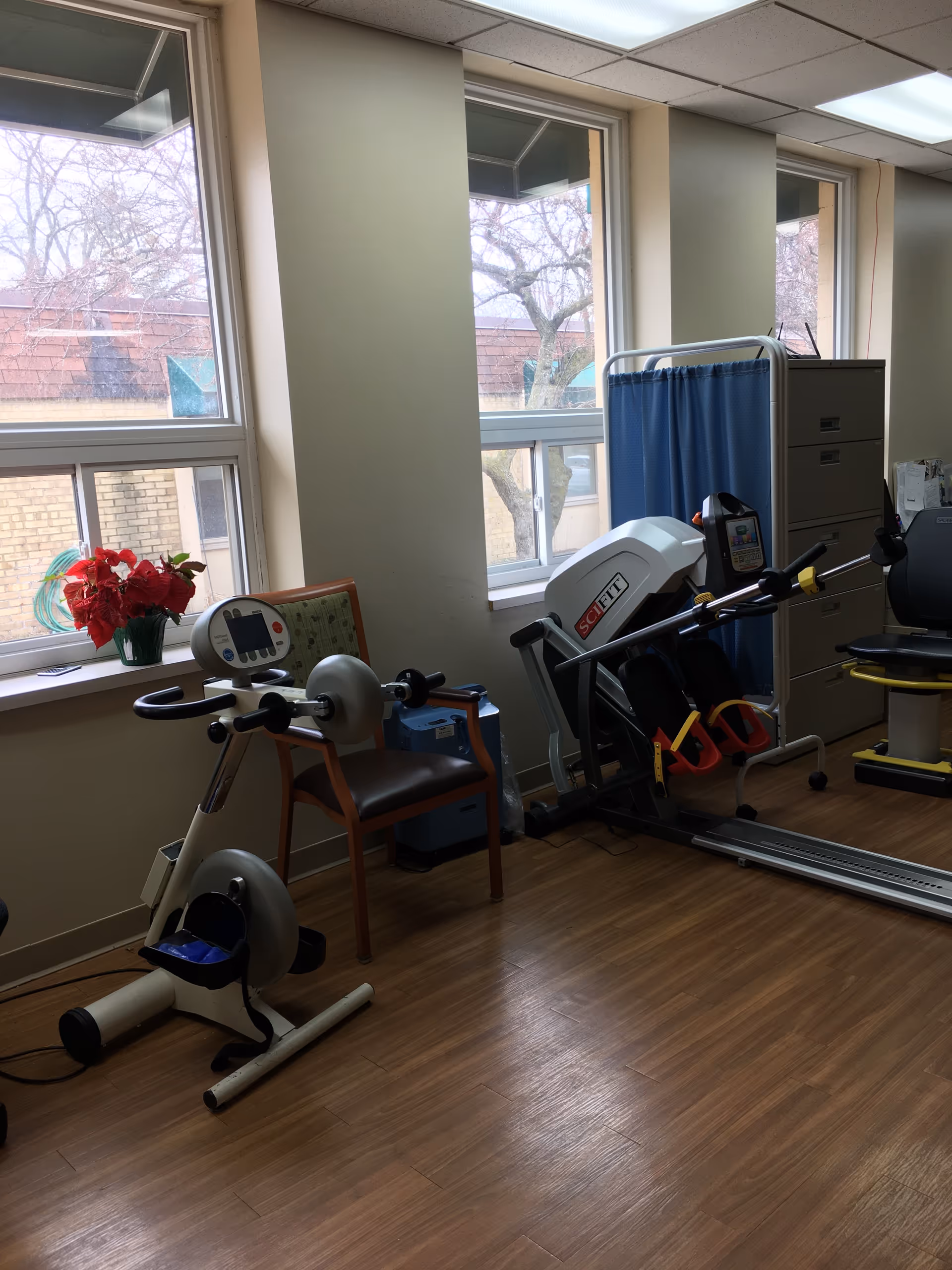 Interior rehab gym with exercise bikes, treadmill, and equipment beside large windows.