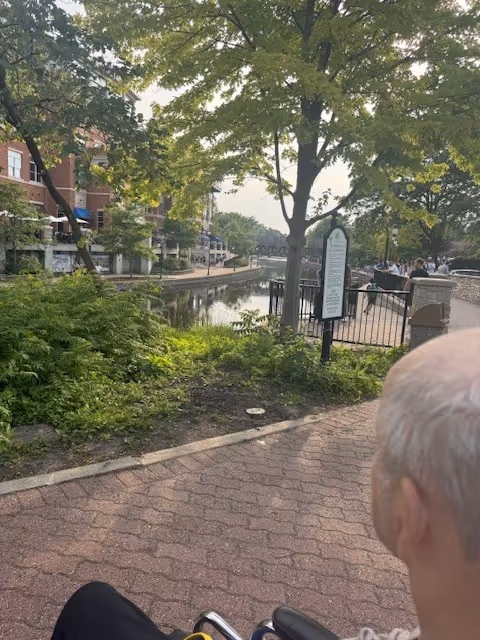 Outdoor canal walkway with trees, railing, signs, and a partial elderly person in foreground.