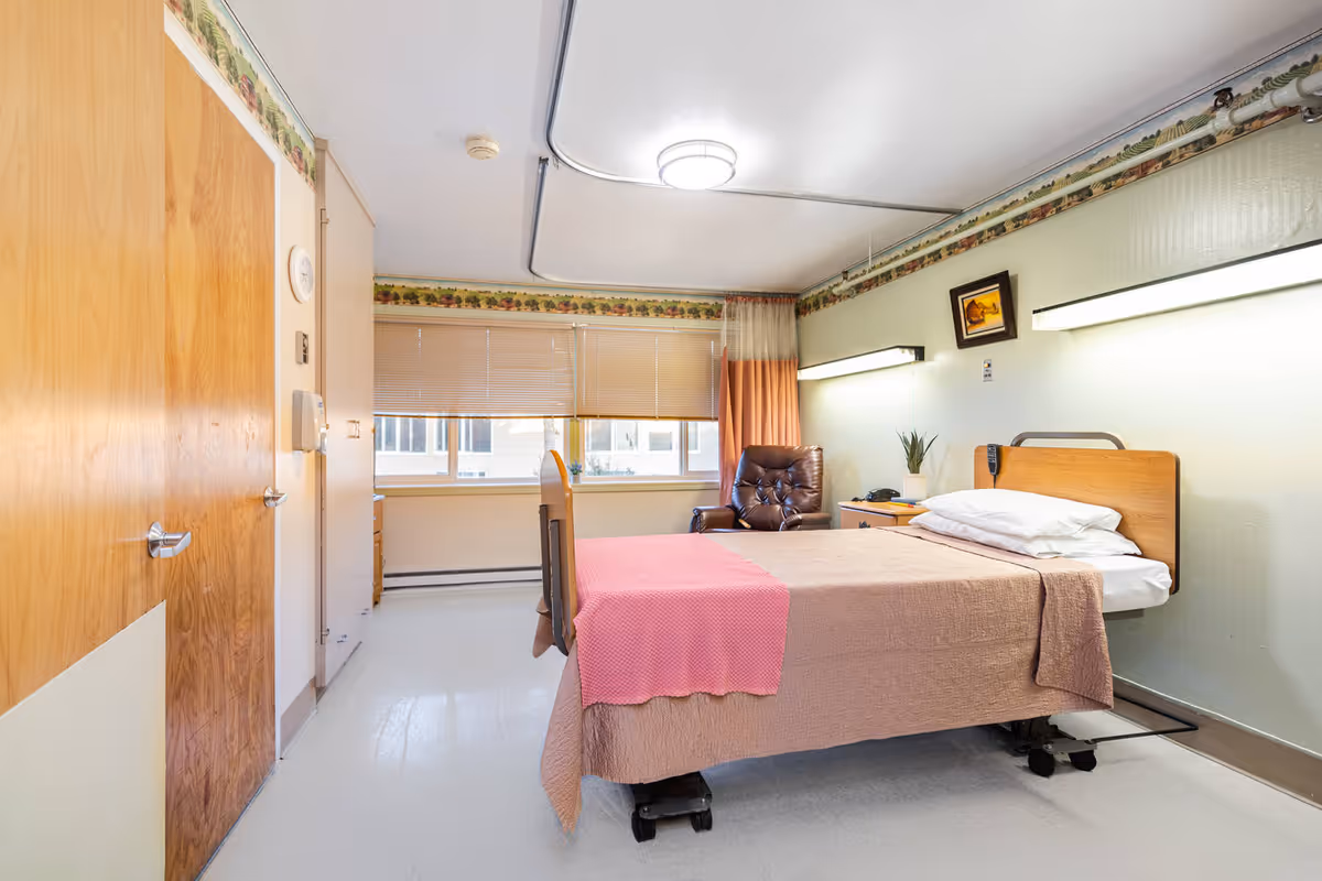 Care facility bedroom with a pink-covered bed, chair, and window blinds.