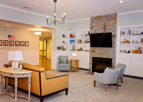 Bright senior living lounge with sofa, chairs, fireplace, built-in shelves, and a wall-mounted TV.
