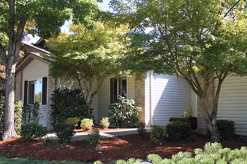 Exterior view of a single-story residential building with white siding and a small porch area, surrounded by green trees and well-maintained landscaping with bushes and mulch.