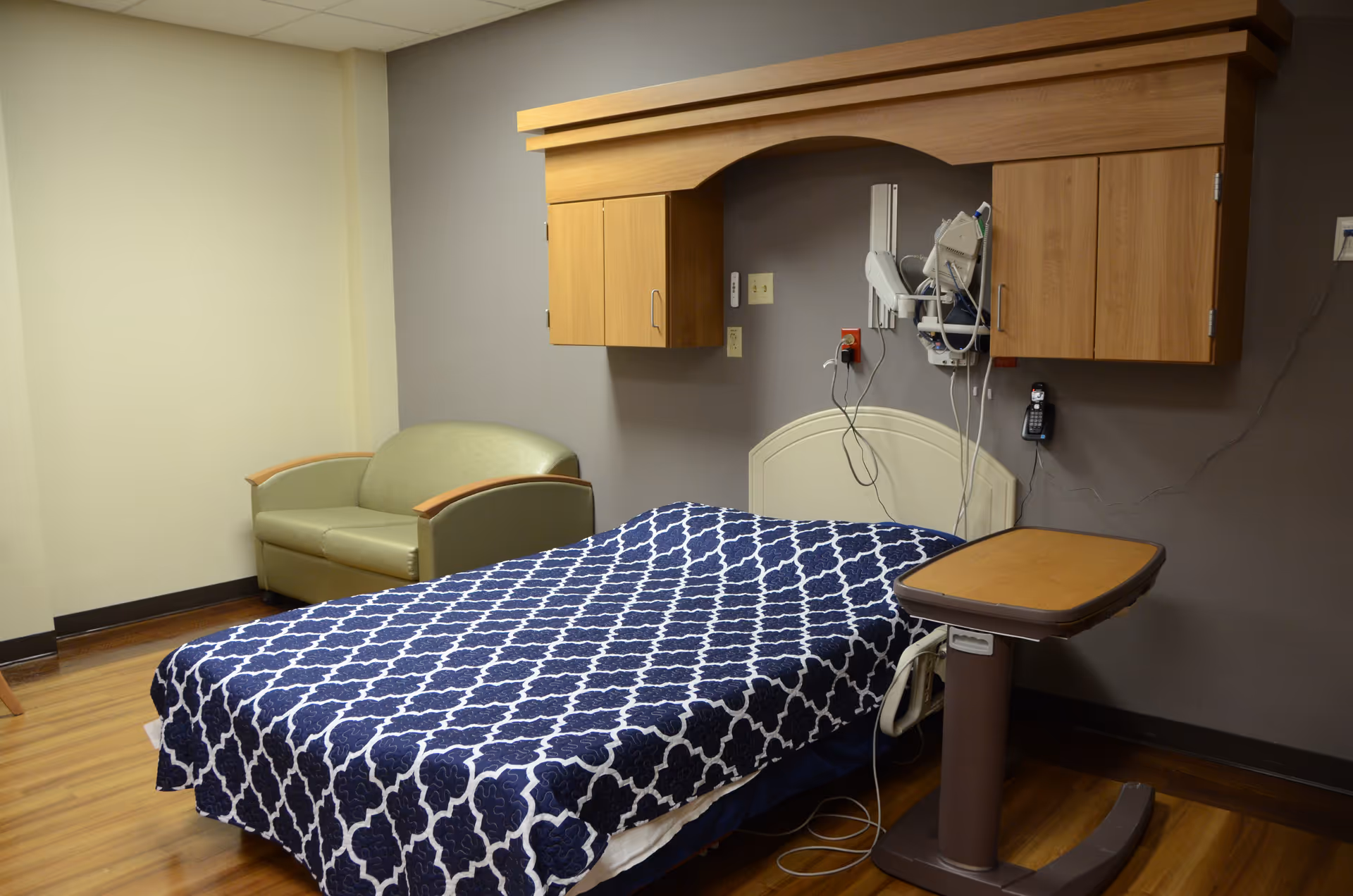 Bedroom in a senior living facility with a patterned bedspread, seating area, and medical equipment.