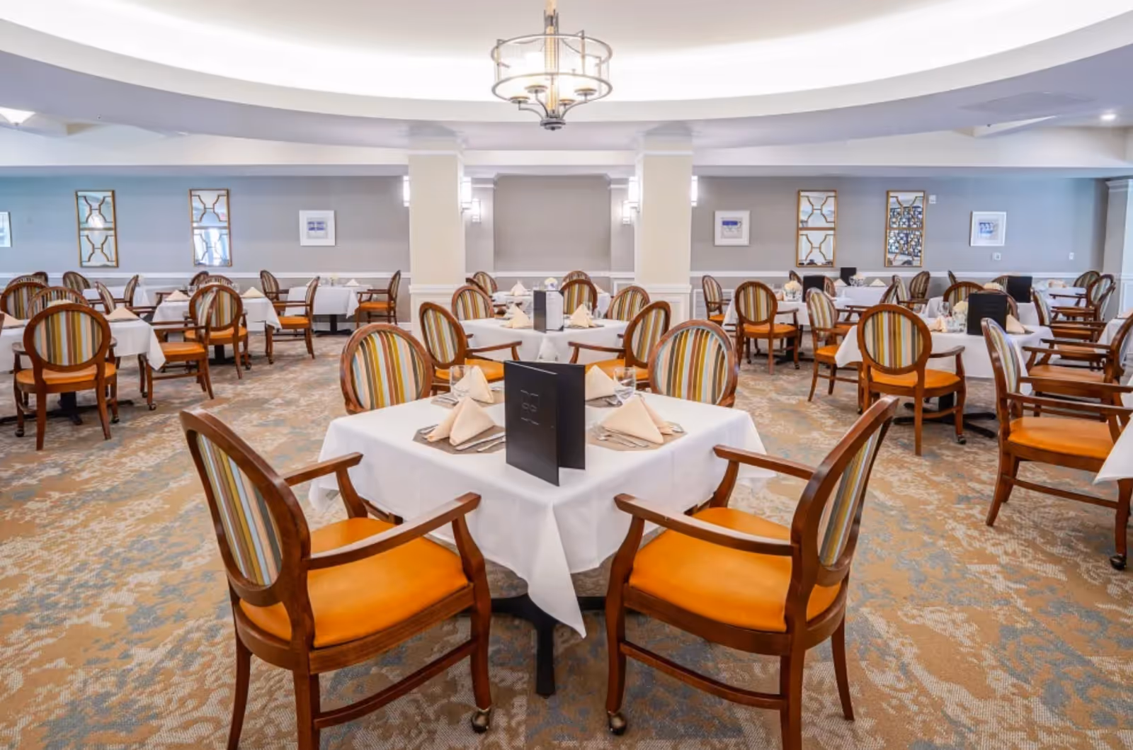 Spacious dining room with tables set with white tablecloths and striped upholstered chairs beneath a central chandelier.