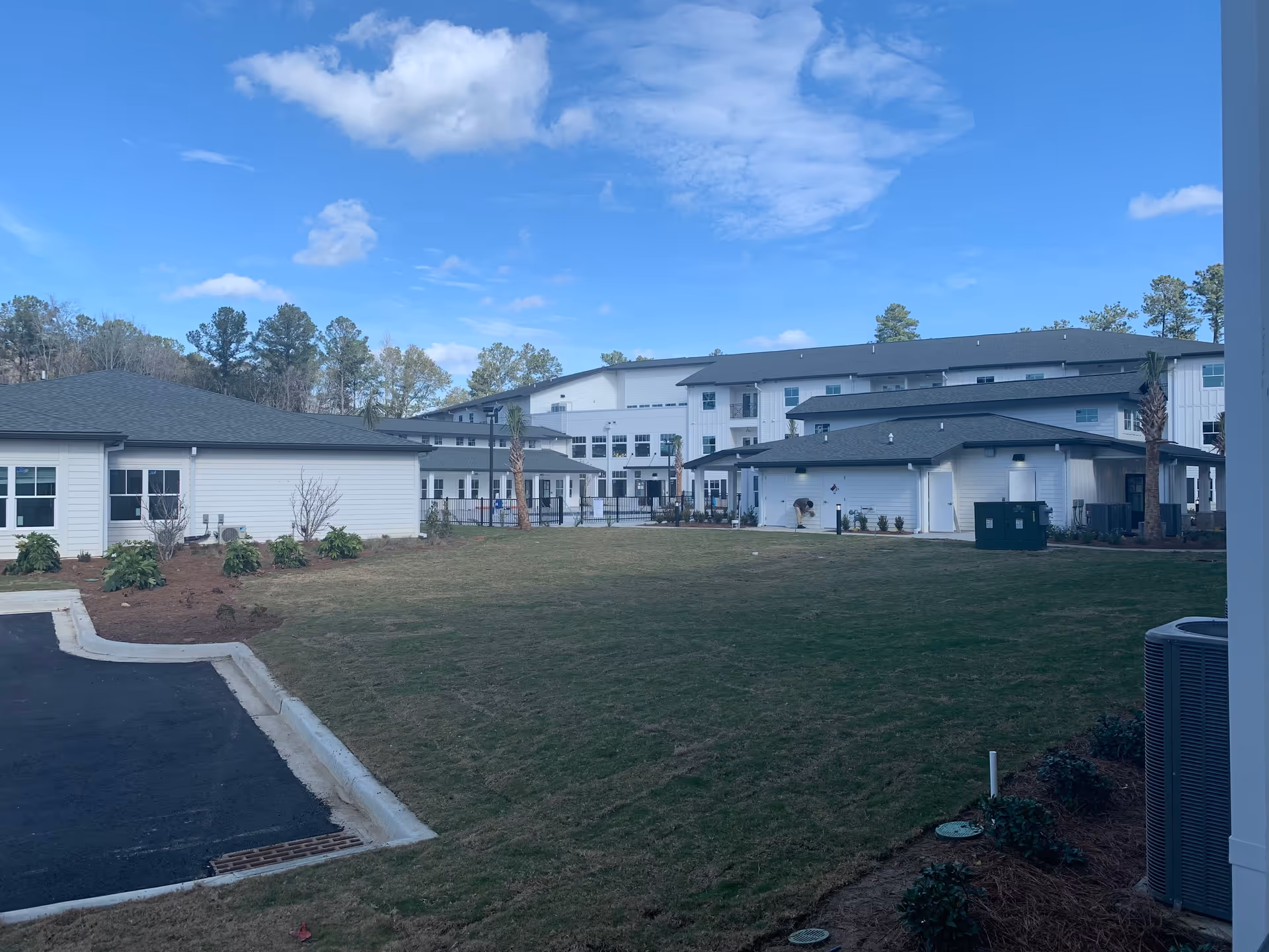 Exterior view of a senior living facility with multiple white buildings featuring dark roofs, surrounded by a grassy lawn and some landscaping. The sky is partly cloudy with blue patches.