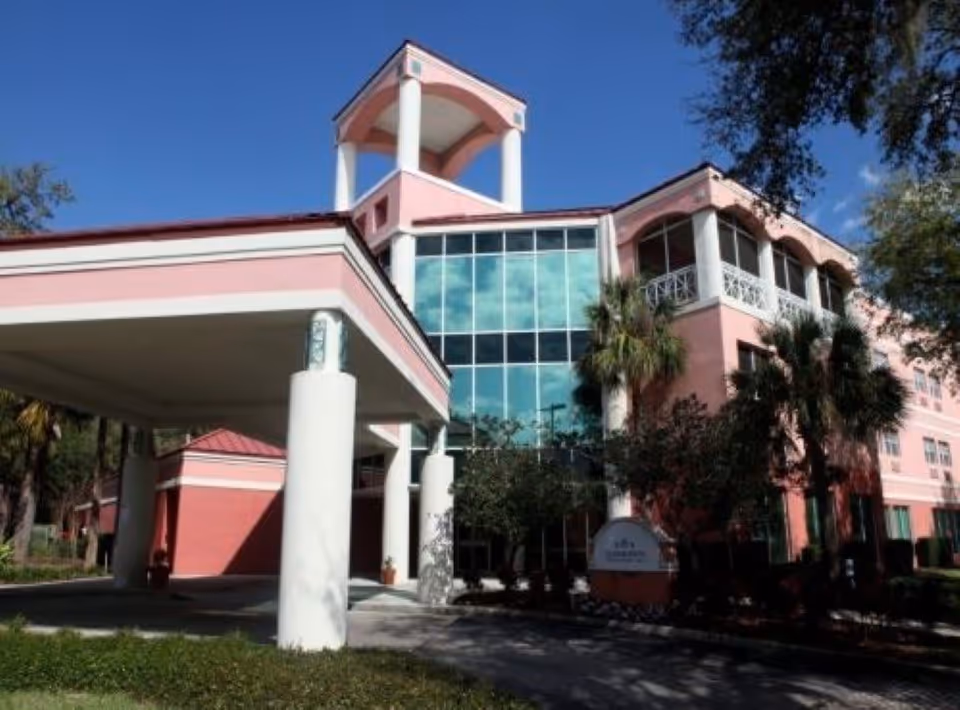 Exterior view of a pink multi-story building with large glass windows and white columns at the entrance, surrounded by trees and greenery under a clear blue sky.