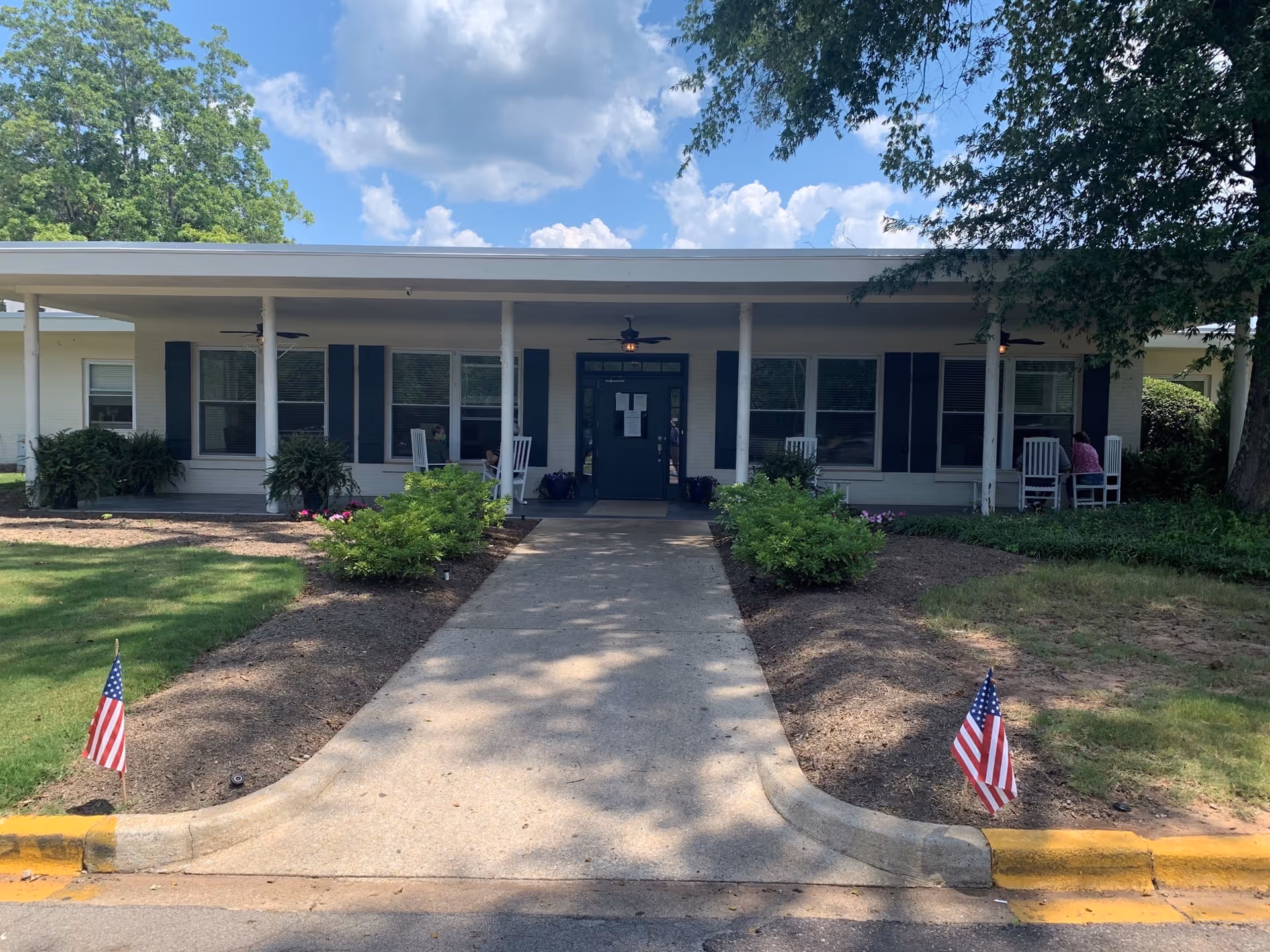 Single-story senior living building with a covered entrance, rocking chairs, and American flags along the walkway.