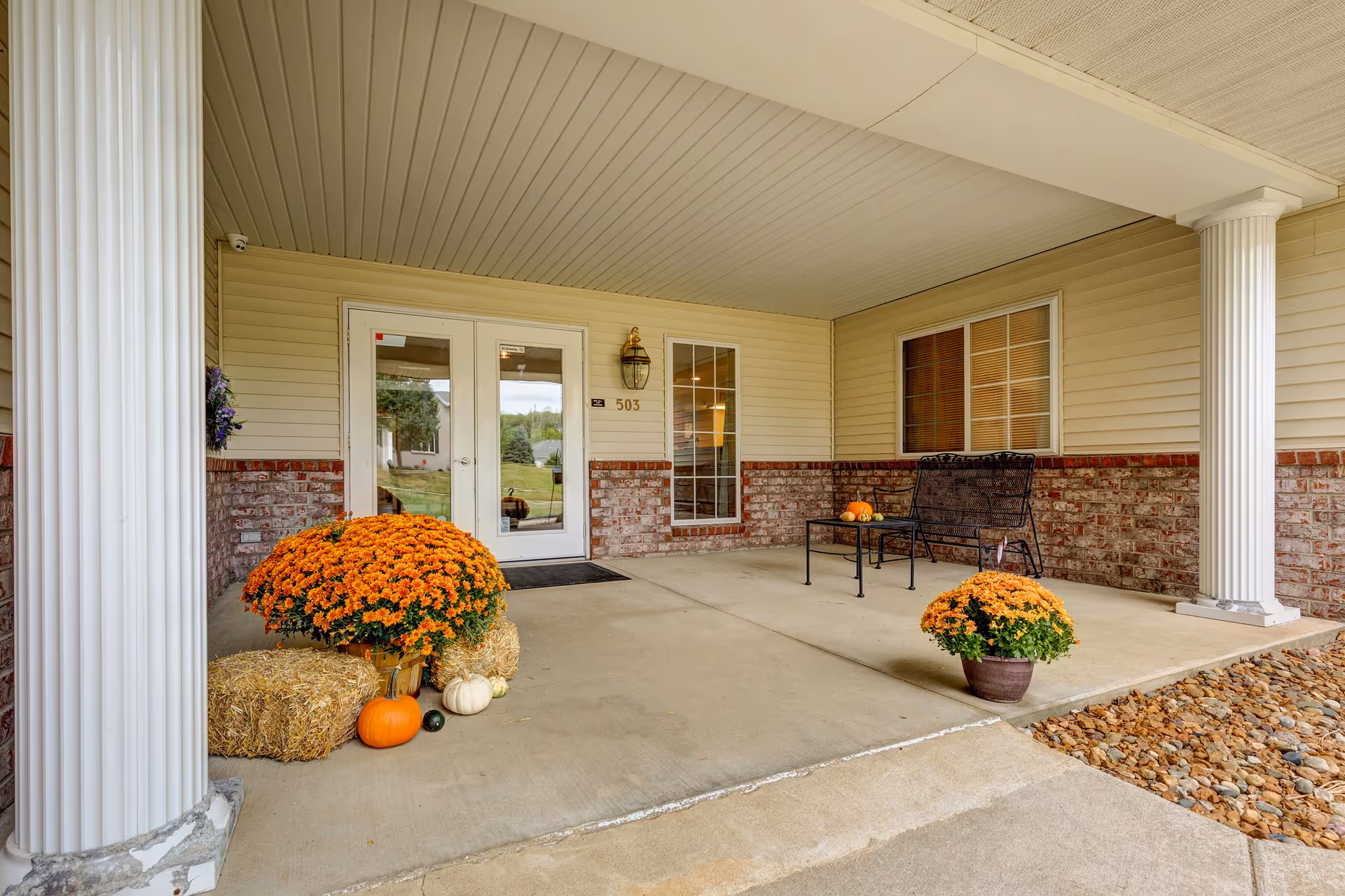 Covered front porch of Shepherd Premier Senior Living with fall pumpkins, mums, hay bales, and seating.