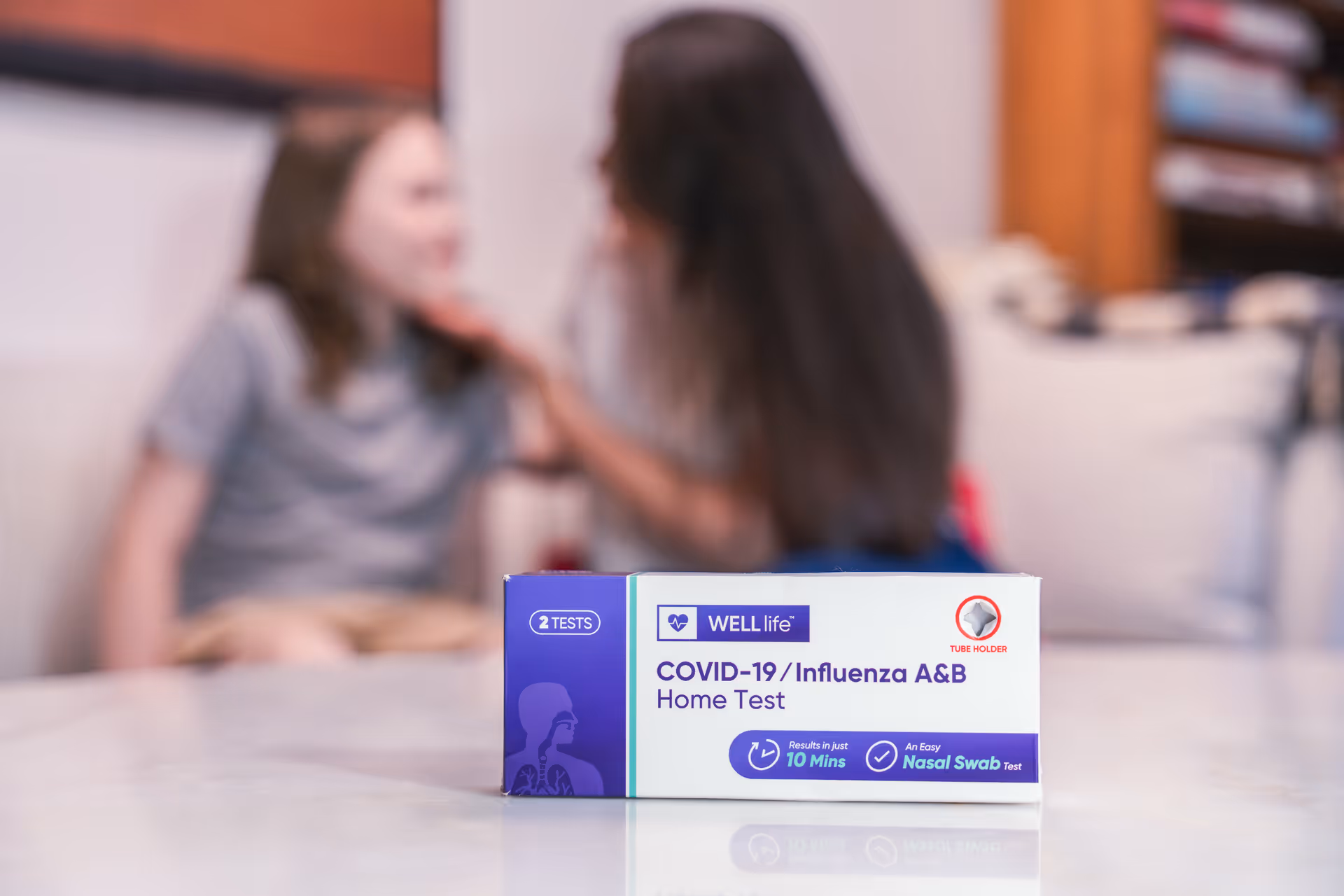 A COVID-19/Influenza home test kit sits on a table with blurred people in background.