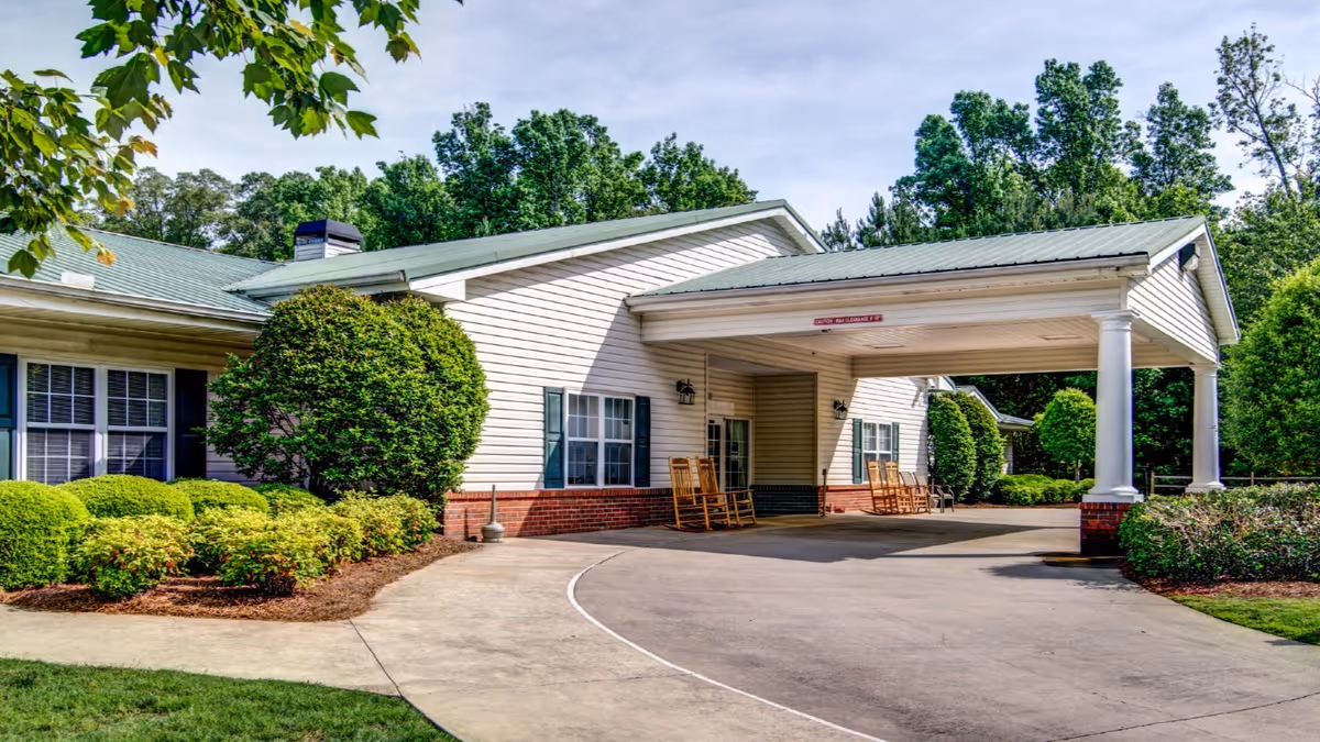 Single-story senior living facility front entrance with a covered porte-cochère, rocking chairs, and manicured landscaping.