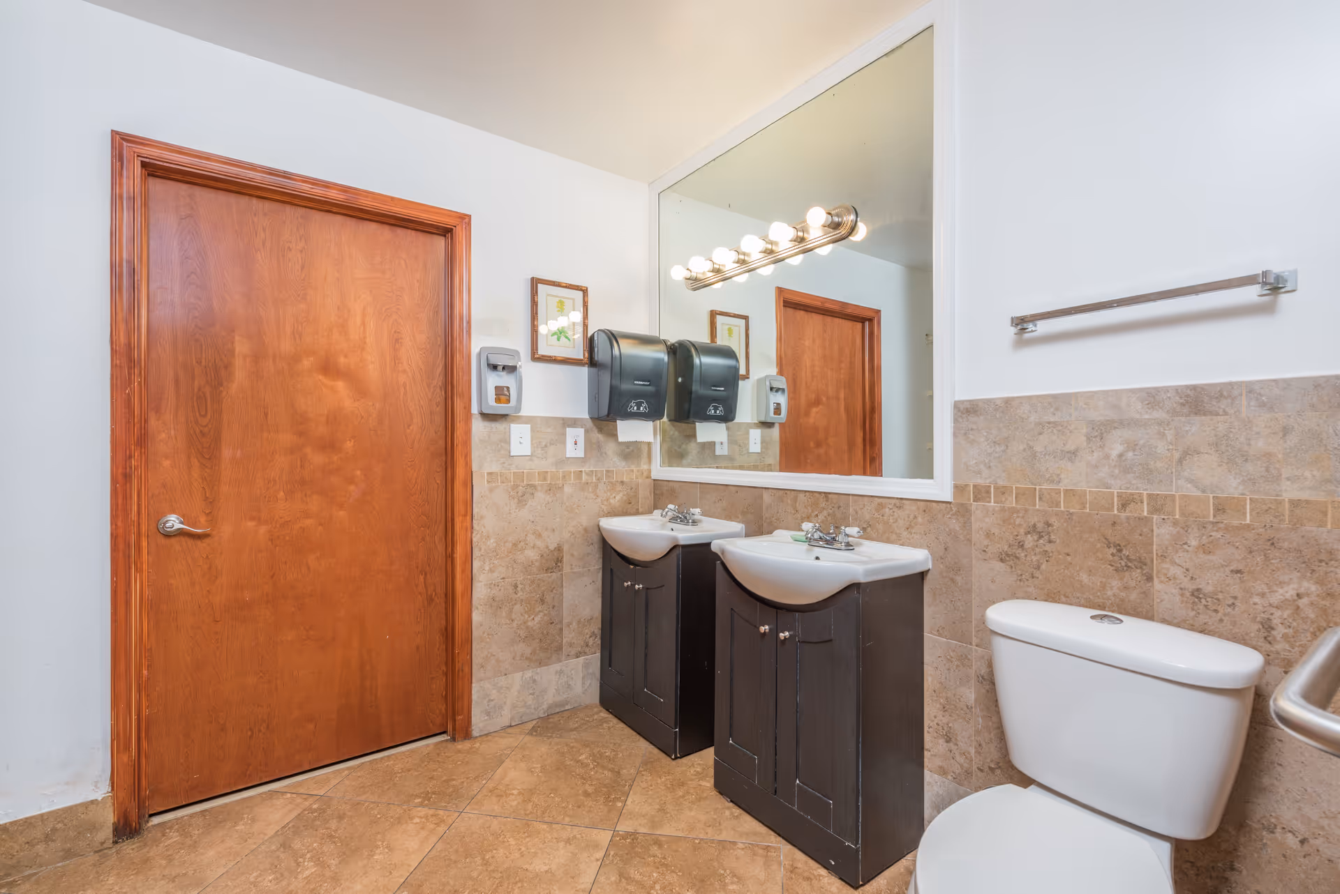 Two sinks with a large mirror, soap dispensers, and a toilet in a tiled bathroom.