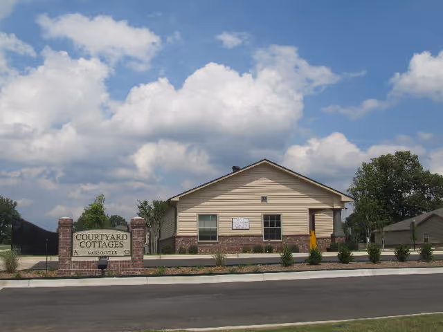 Front exterior of a single-story Courtyard Cottages building with an entrance sign, low shrubs, and a partly cloudy sky.