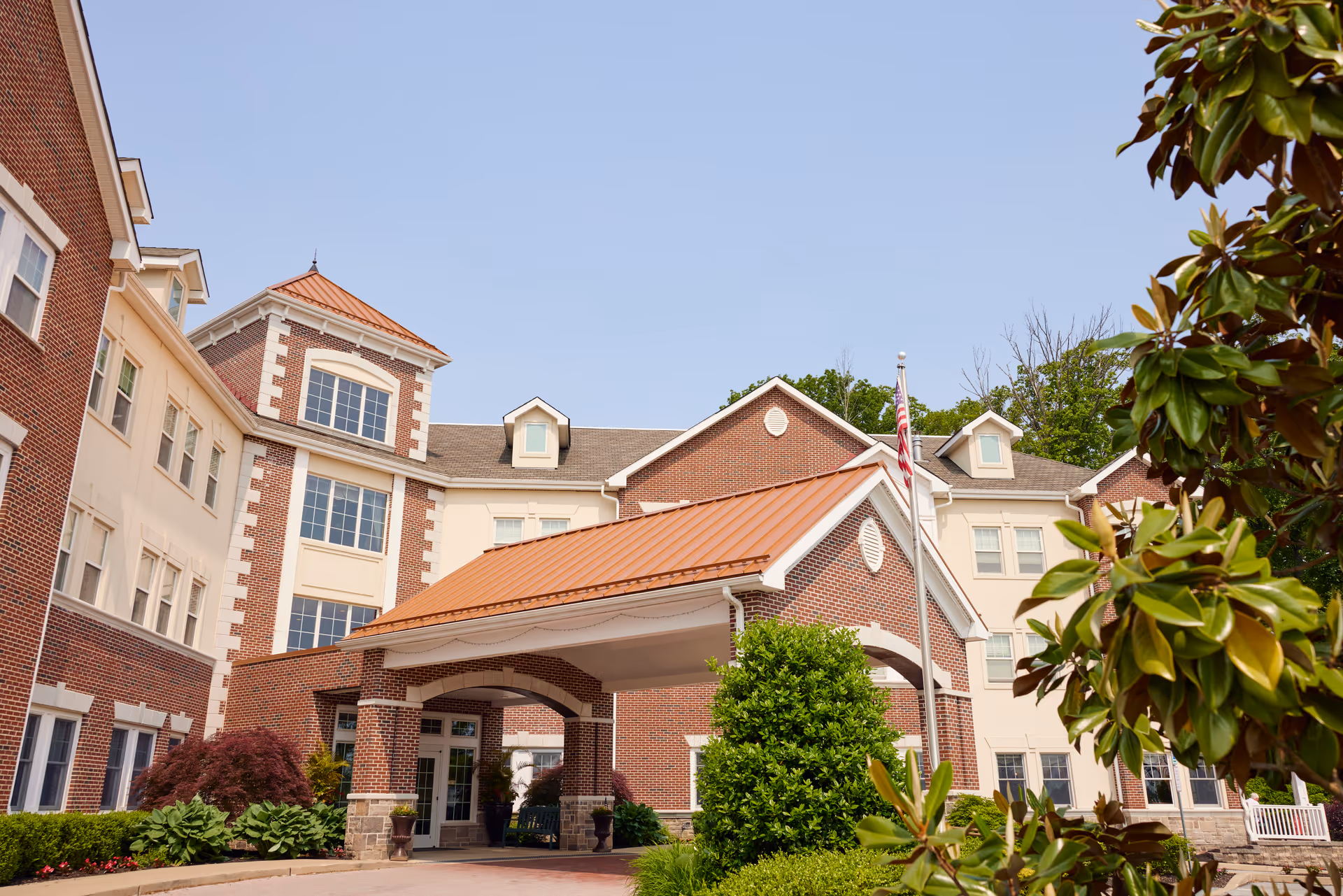 Front exterior of a multi-story brick senior living building with a covered porte-cochère entrance and surrounding landscaping.