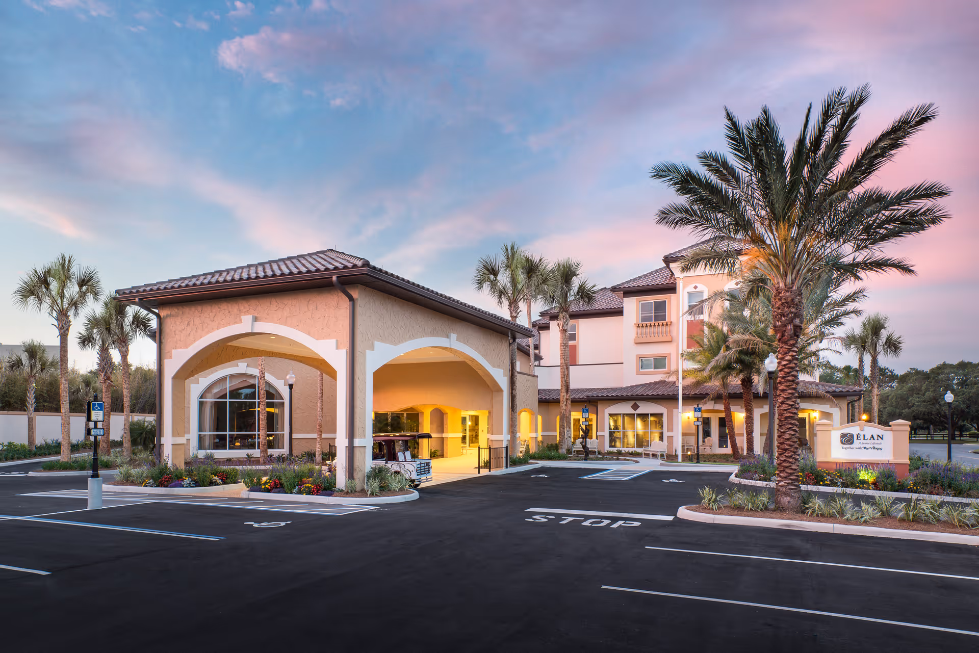 Front exterior of a senior living building with arched entry, palm trees, and a parking lot at sunset.