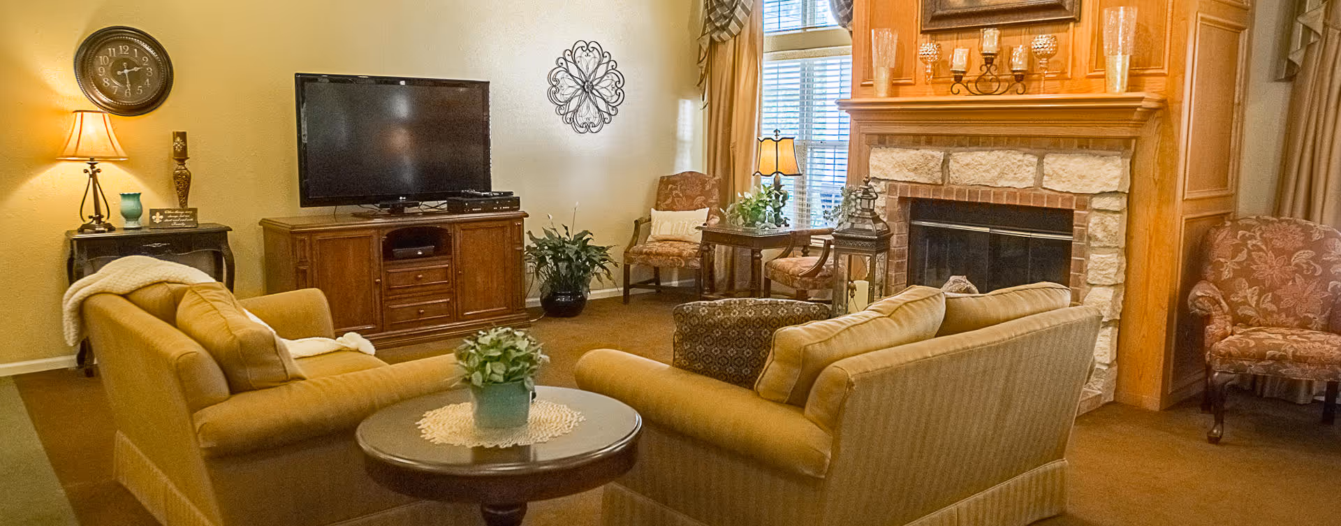 A cozy living room with two beige sofas facing a flat-screen TV on a wooden cabinet. There is a round wooden table with a small plant in the center, a stone fireplace with candles on the mantel, and several upholstered chairs. The room is warmly lit with a table lamp and natural light coming through a window with curtains.