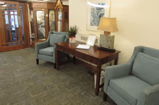 A cozy interior seating area with two blue upholstered armchairs on either side of a wooden console table. The table holds a lamp with a beige shade, a small plant, and some papers. Behind the seating area is a wooden grandfather clock and a glass-paneled door.