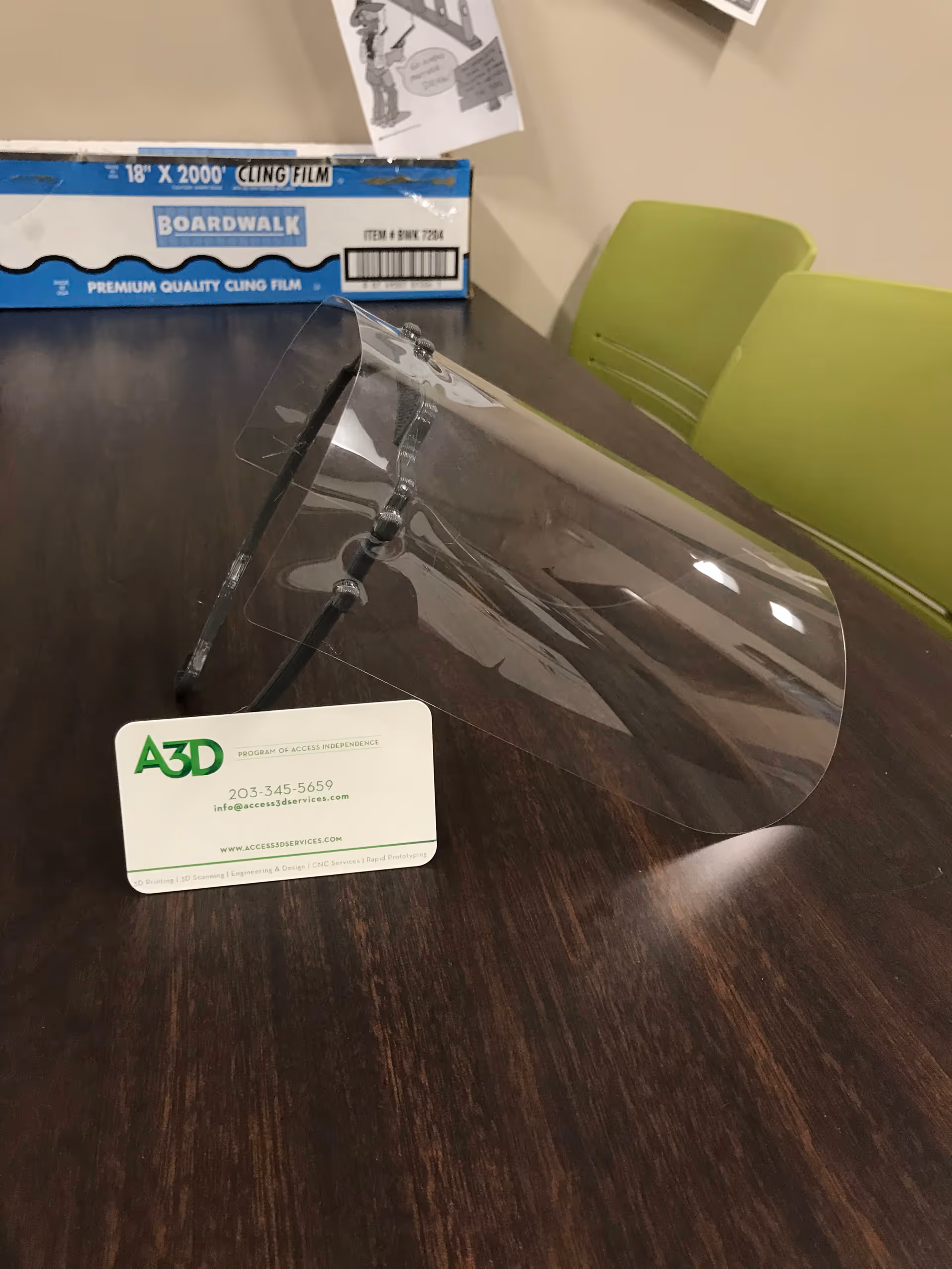 Transparent face shield and a business card on a dark wooden table.