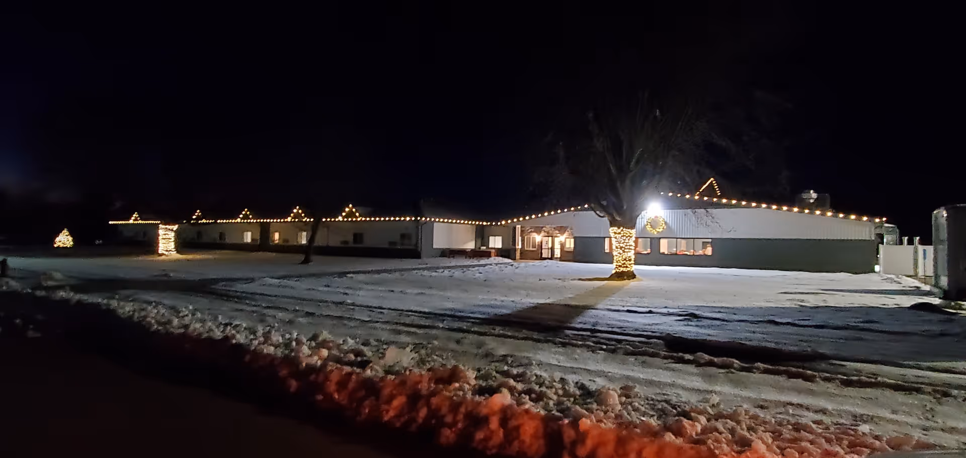 Night view of a single-story facility building decorated with string lights and a tree wrapped in lights, with snow-covered lawn in the foreground.