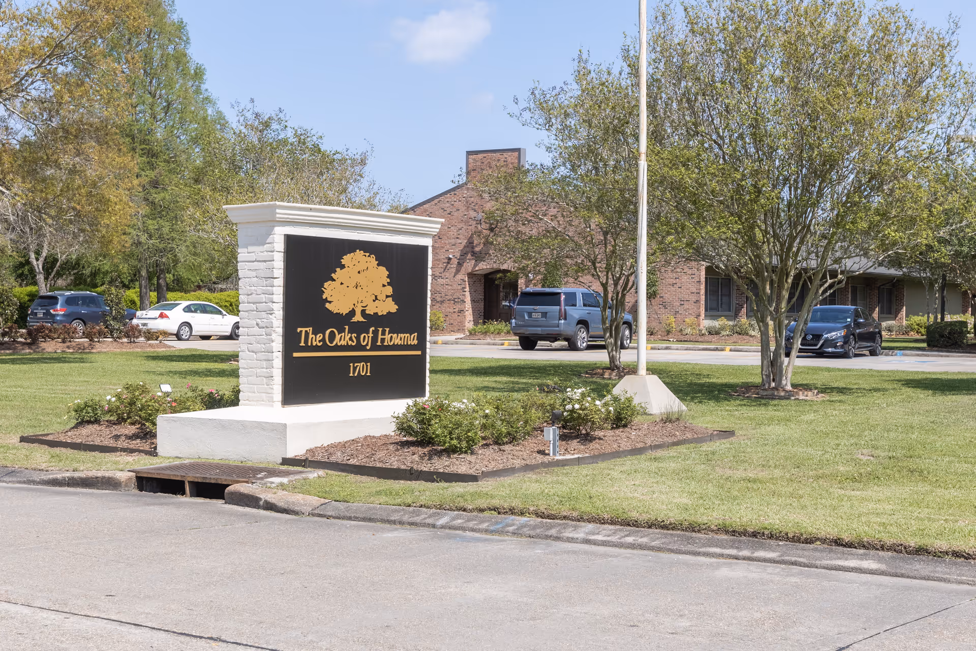 Outdoor view of The Oaks of Houma senior living facility sign with a large oak tree logo and address number 1701. The sign is situated on a landscaped grassy area with trees and bushes, and a brick building with parked cars is visible in the background under a clear blue sky.