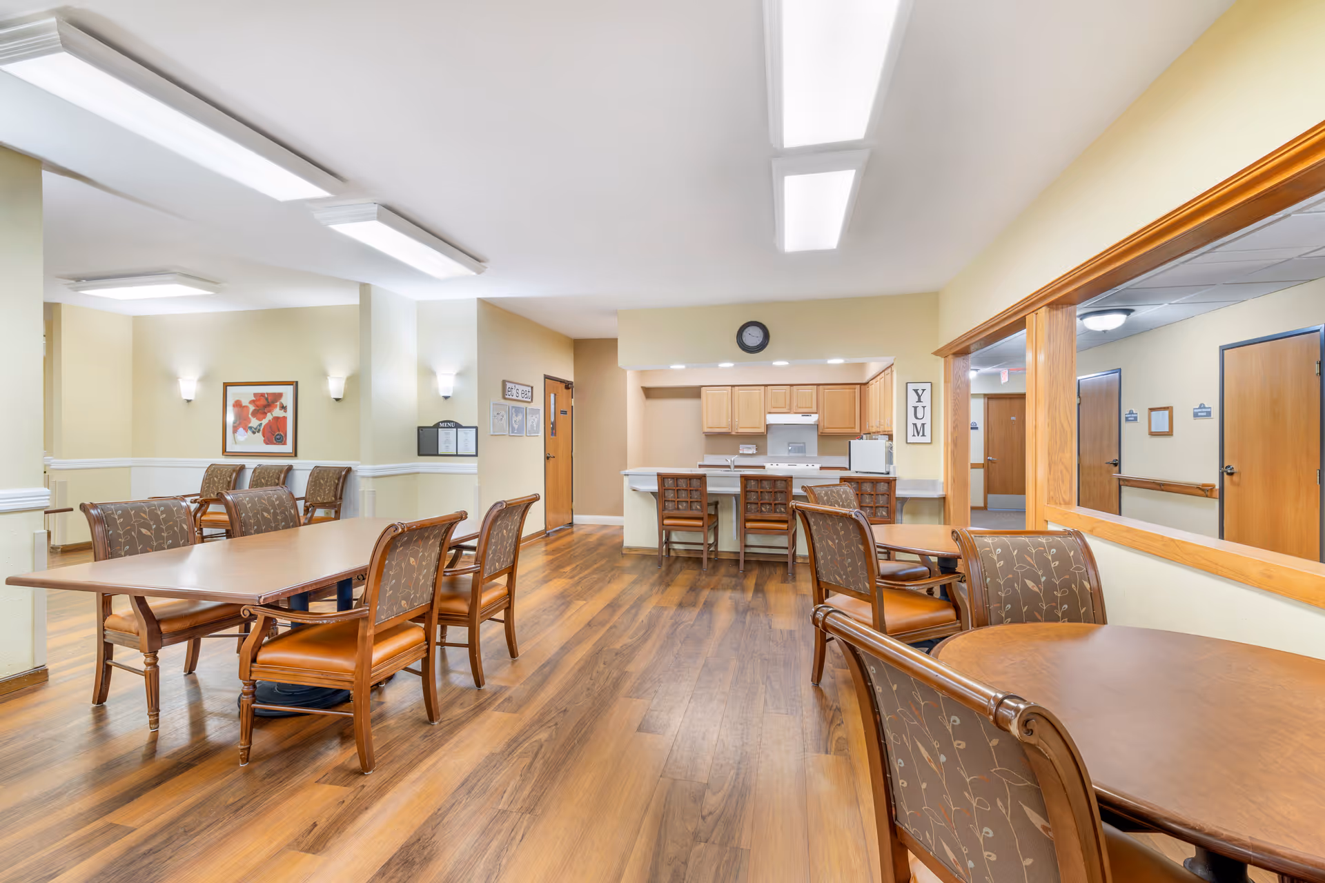 Bright communal dining area with wooden tables, chairs, and a service counter in the background.