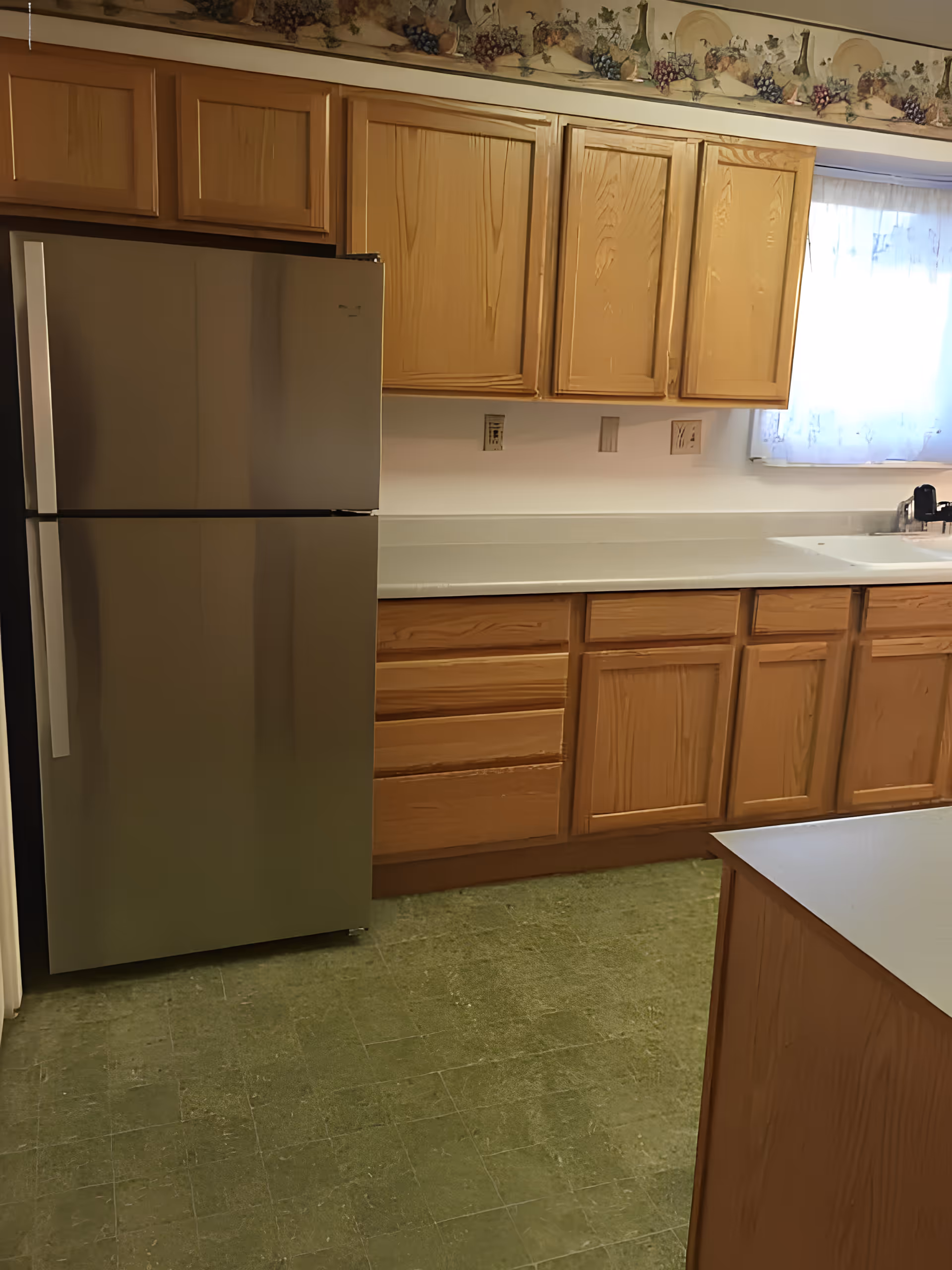 A kitchen with wooden cabinets, a stainless steel refrigerator, a white countertop, and a window with a sheer curtain. The floor is covered with green linoleum tiles and there is a decorative border with grapes and leaves along the top of the wall.