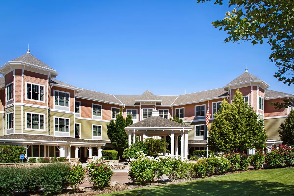 Three-story senior living building with a central covered entrance, landscaped flower beds, and an American flag under a clear blue sky.