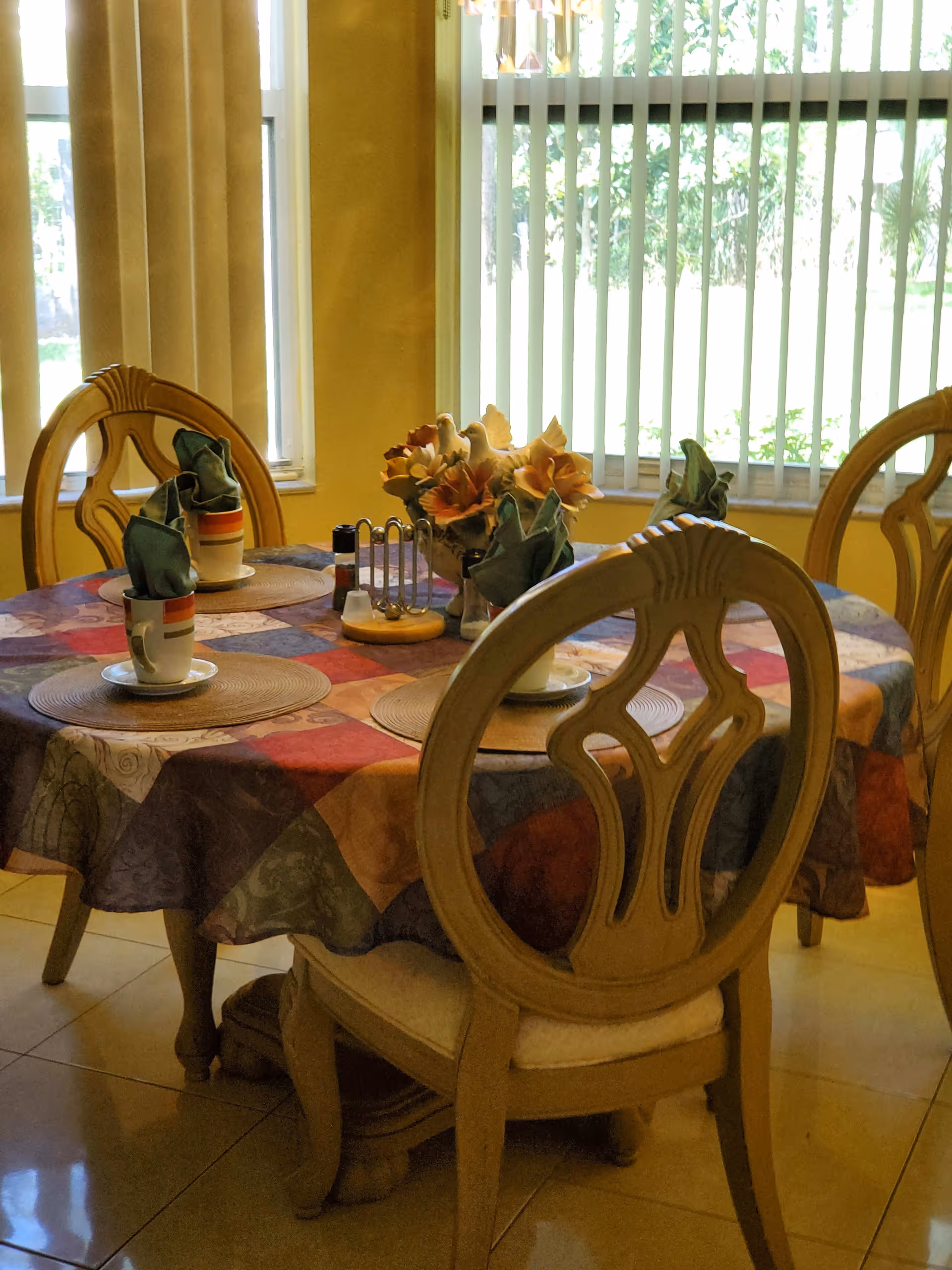 Wooden dining table with a colorful tablecloth, place settings, napkins and a floral centerpiece in front of vertical window blinds.