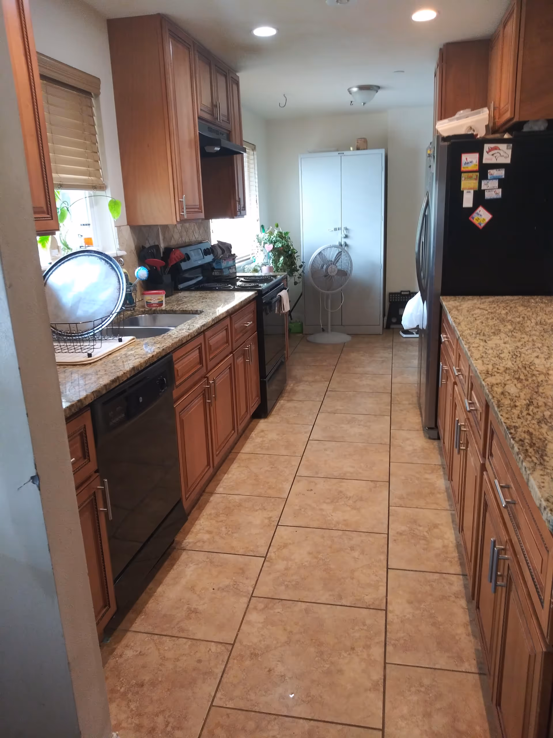 A narrow galley kitchen with brown cabinets, granite counters, appliances, and a standing fan.