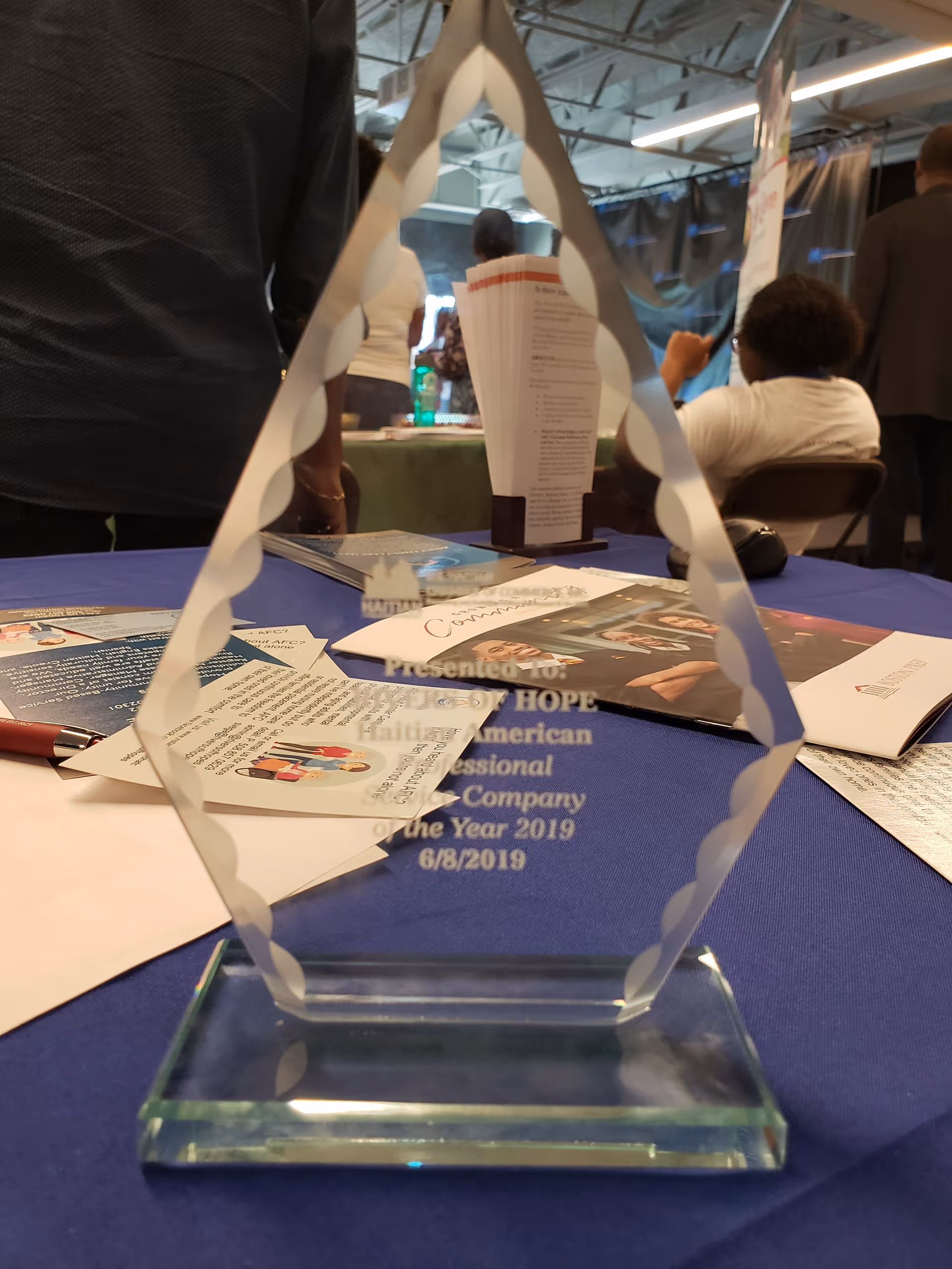 Crystal trophy on a blue table with brochures and people in the background.