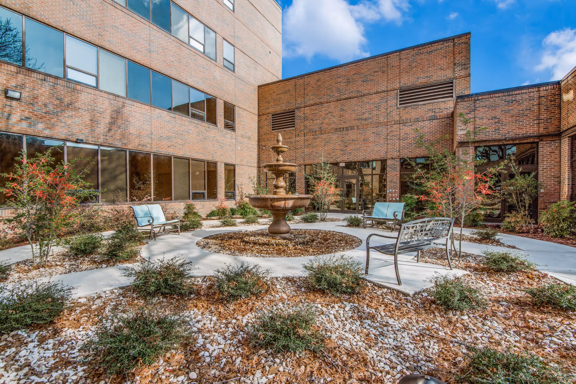 Outdoor courtyard area at The Heritage at Hunters Chase featuring a central three-tiered water fountain surrounded by a circular walkway, benches with light blue cushions, small shrubs, and trees with red berries. The courtyard is enclosed by brick building walls with multiple windows reflecting the blue sky and clouds.