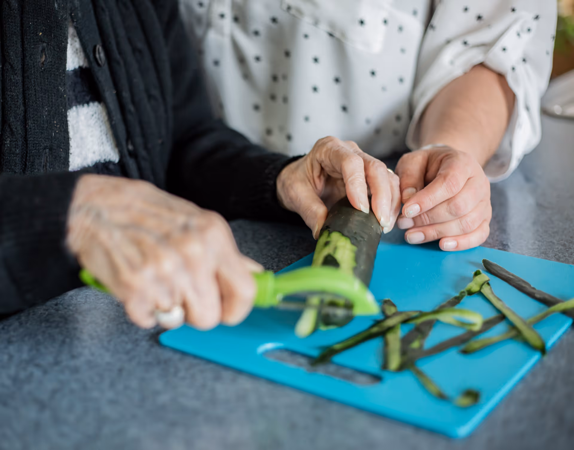 Close-up of elderly hands slicing green vegetables on a blue cutting board, with assistance.