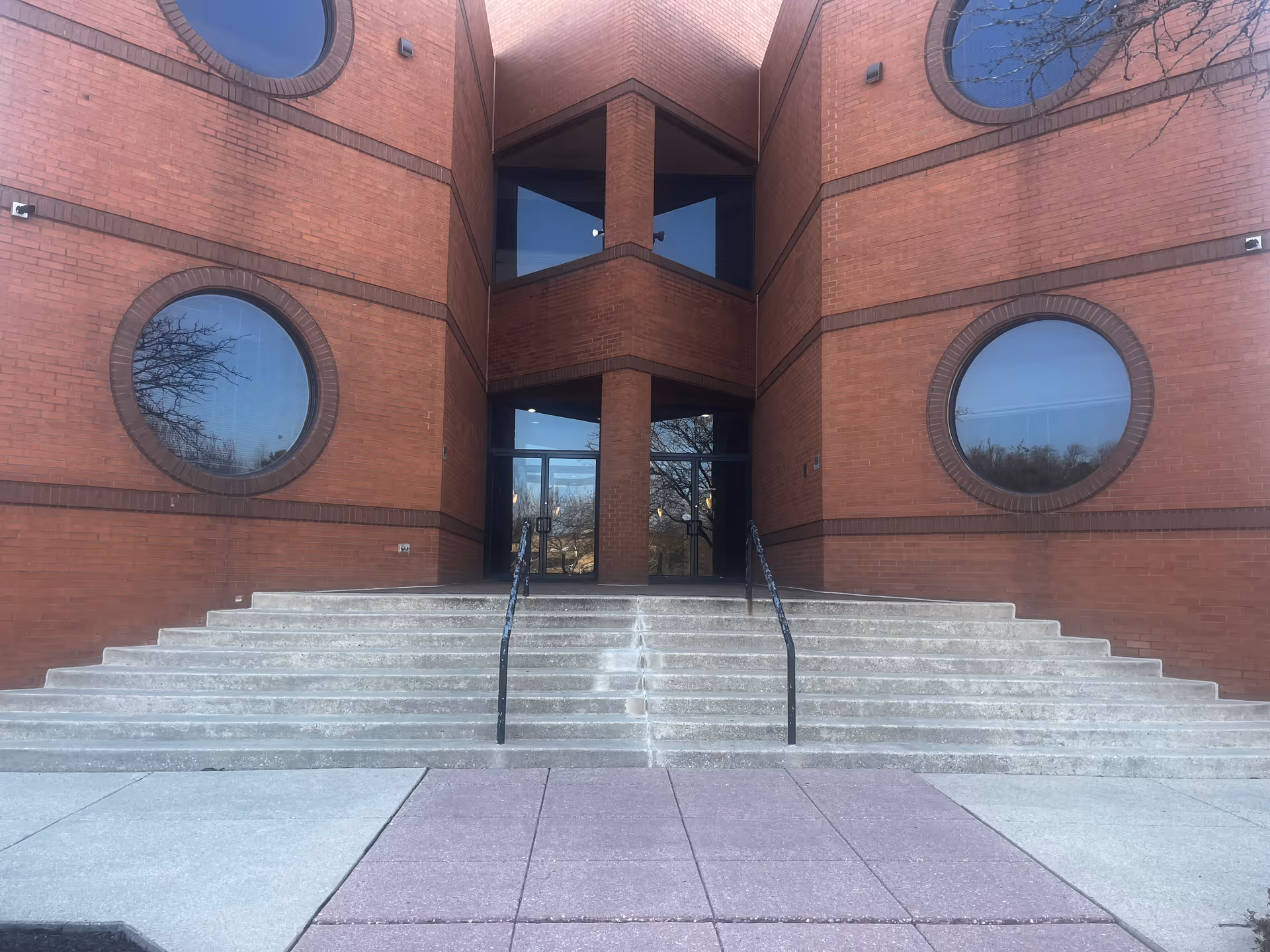Red brick building entrance with circular windows and concrete steps up to glass doors.