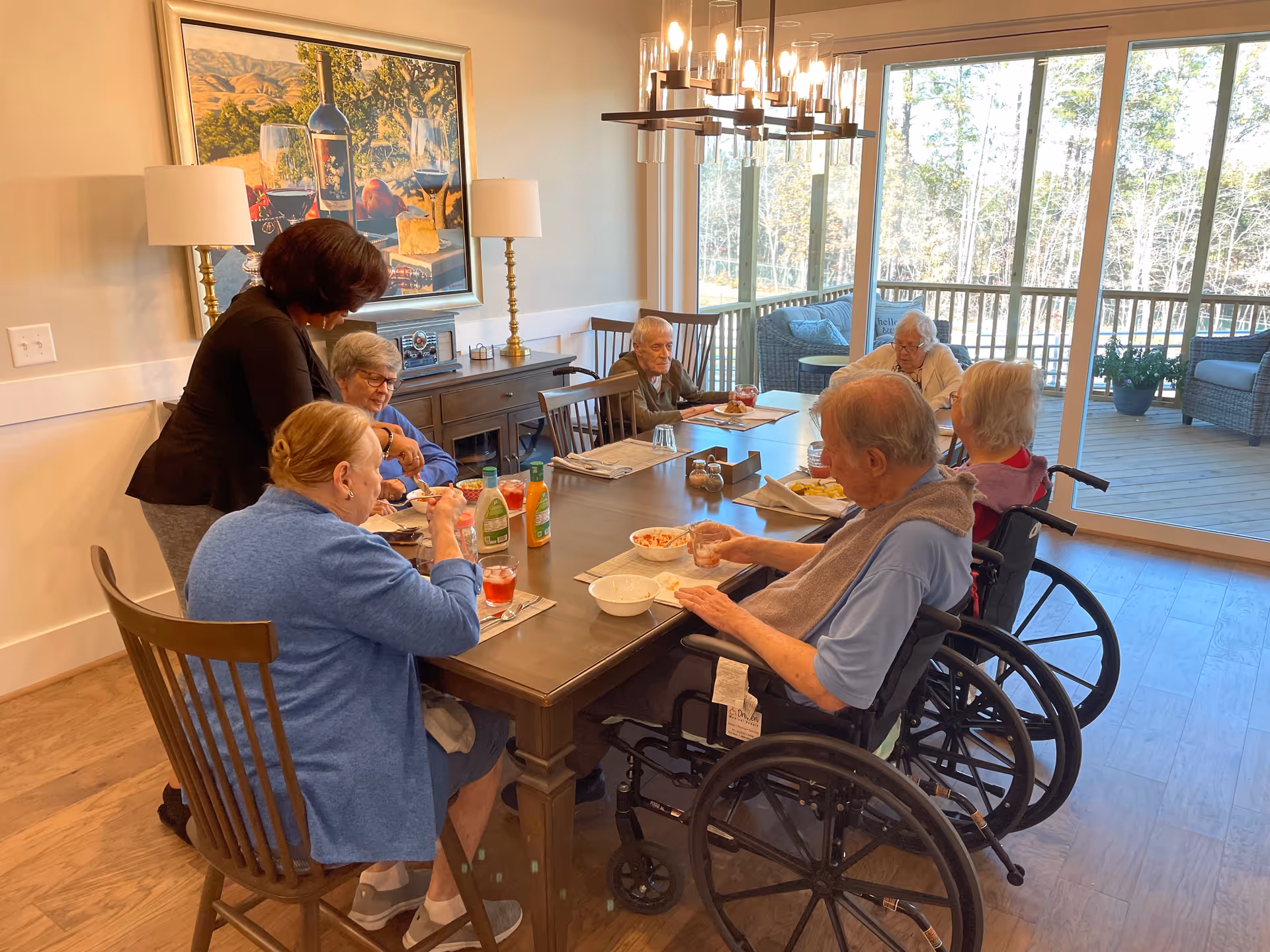Residents, some in wheelchairs, share a meal around a large dining table with a staff member.