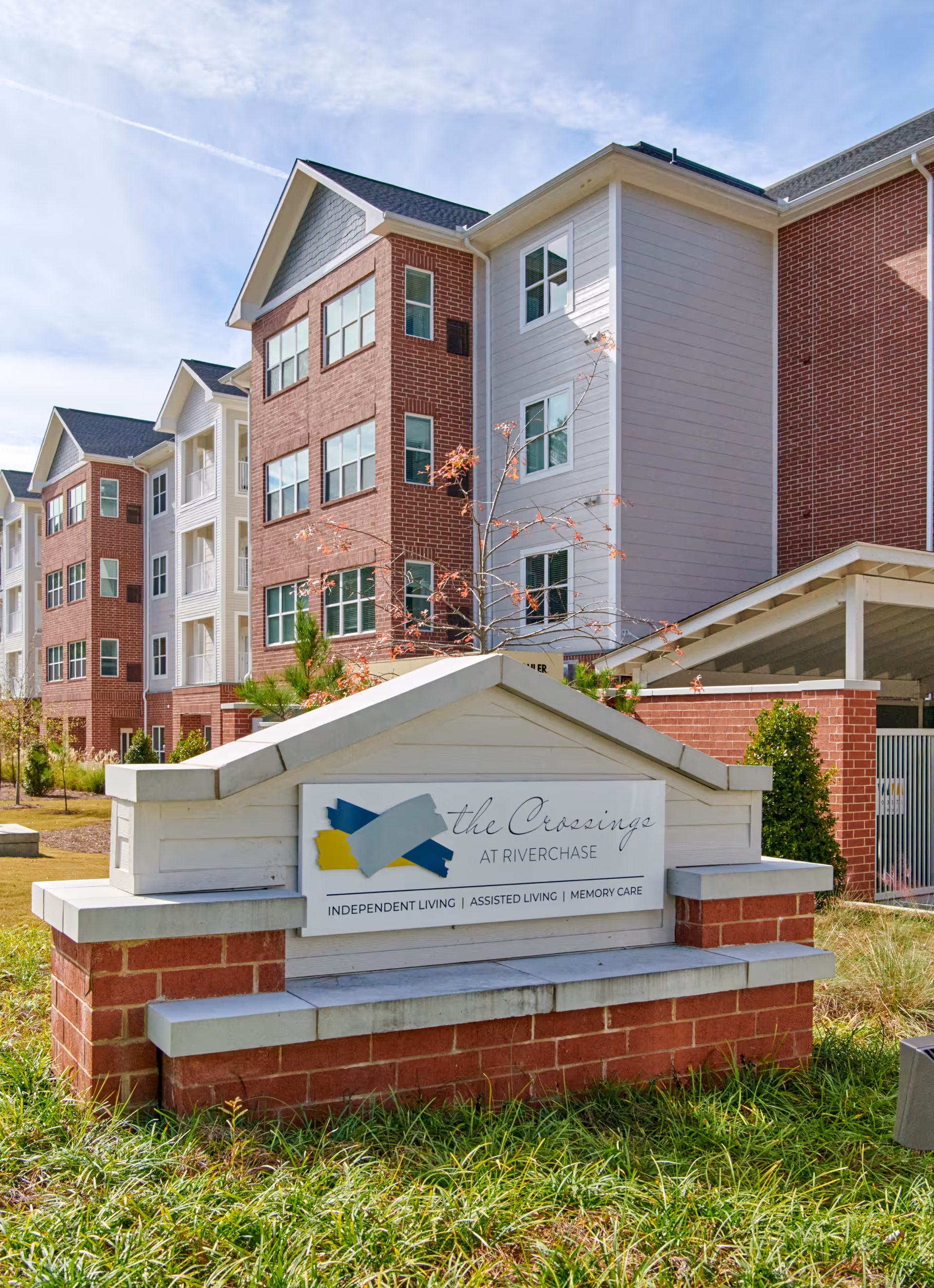 Exterior view of The Crossings at Riverchase senior living facility with a brick and white siding multi-story building in the background and a large sign in the foreground displaying the facility name and services including Independent Living, Assisted Living, and Memory Care.