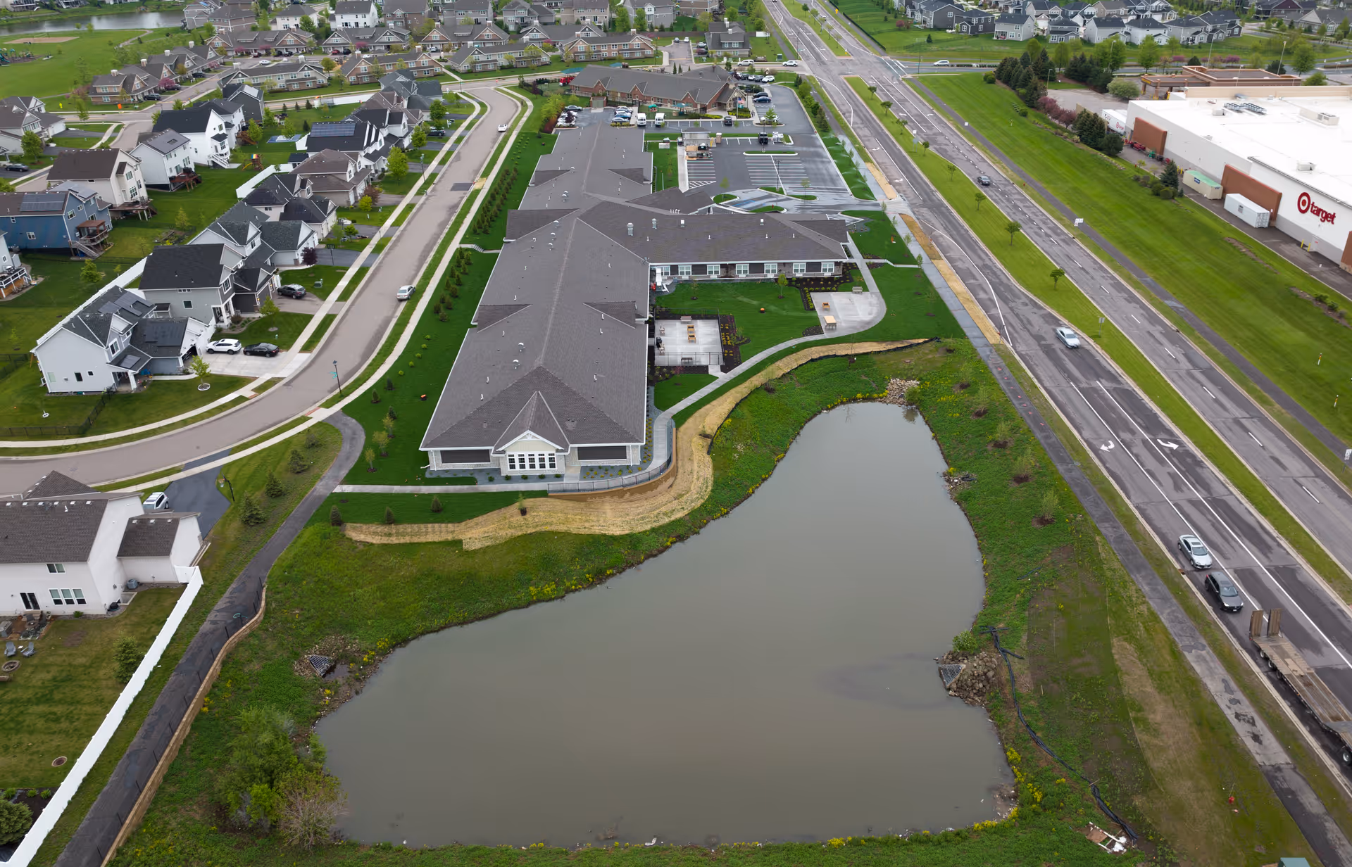 Aerial view of Boden Senior Living facility showing a large building with a dark roof surrounded by green lawns, a small pond in the foreground, a road with cars on the right, and a residential neighborhood with houses on the left.