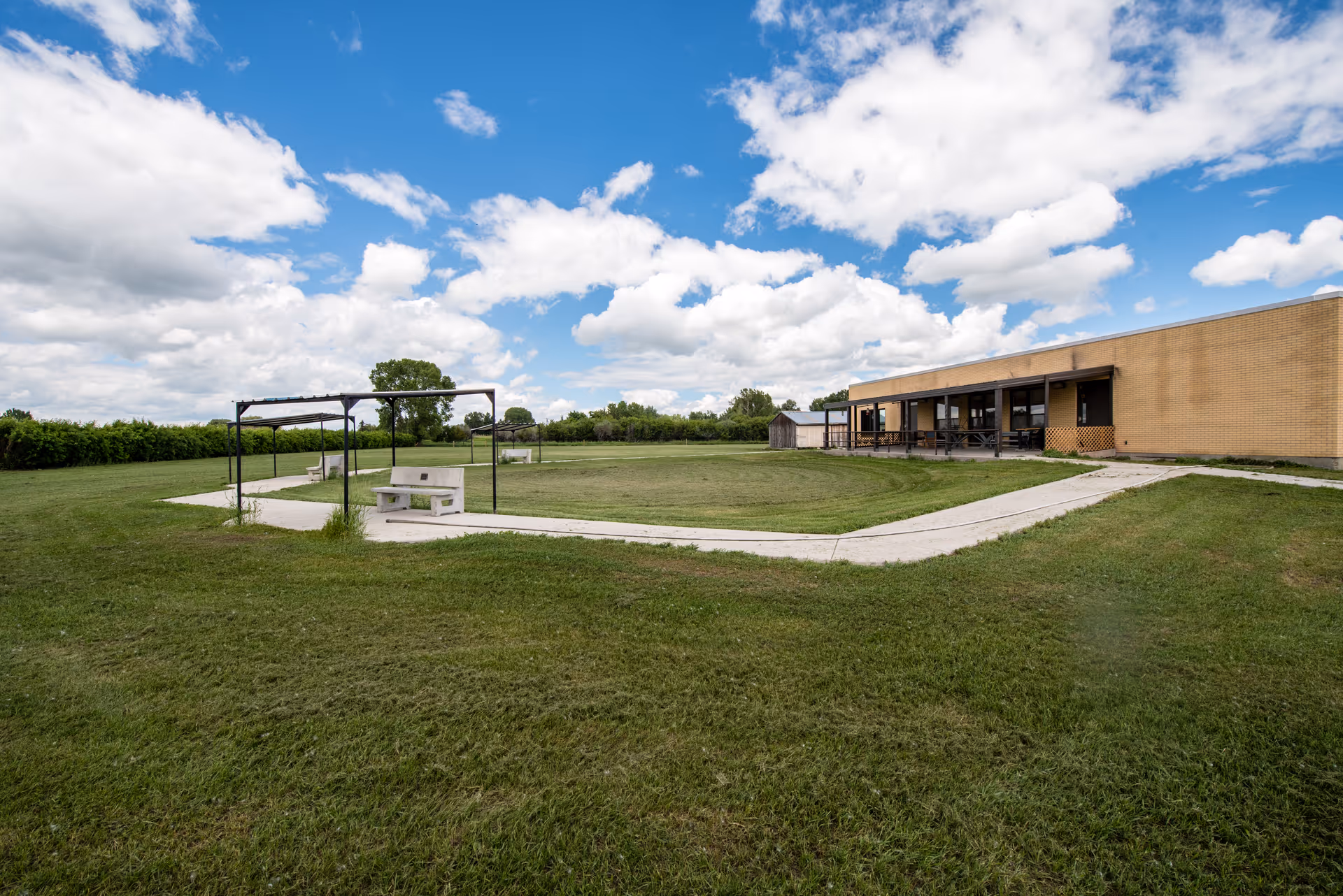 Wide outdoor courtyard with a brick building, concrete paths, and a metal pergola with benches.