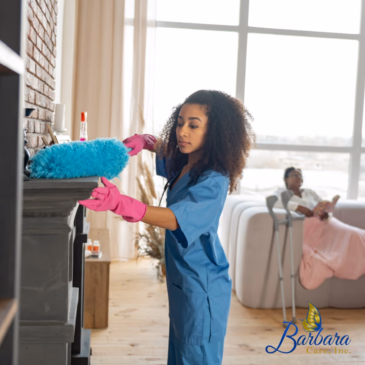 Caregiver in blue scrubs dusts a fireplace while a resident relaxes in a bright living room.